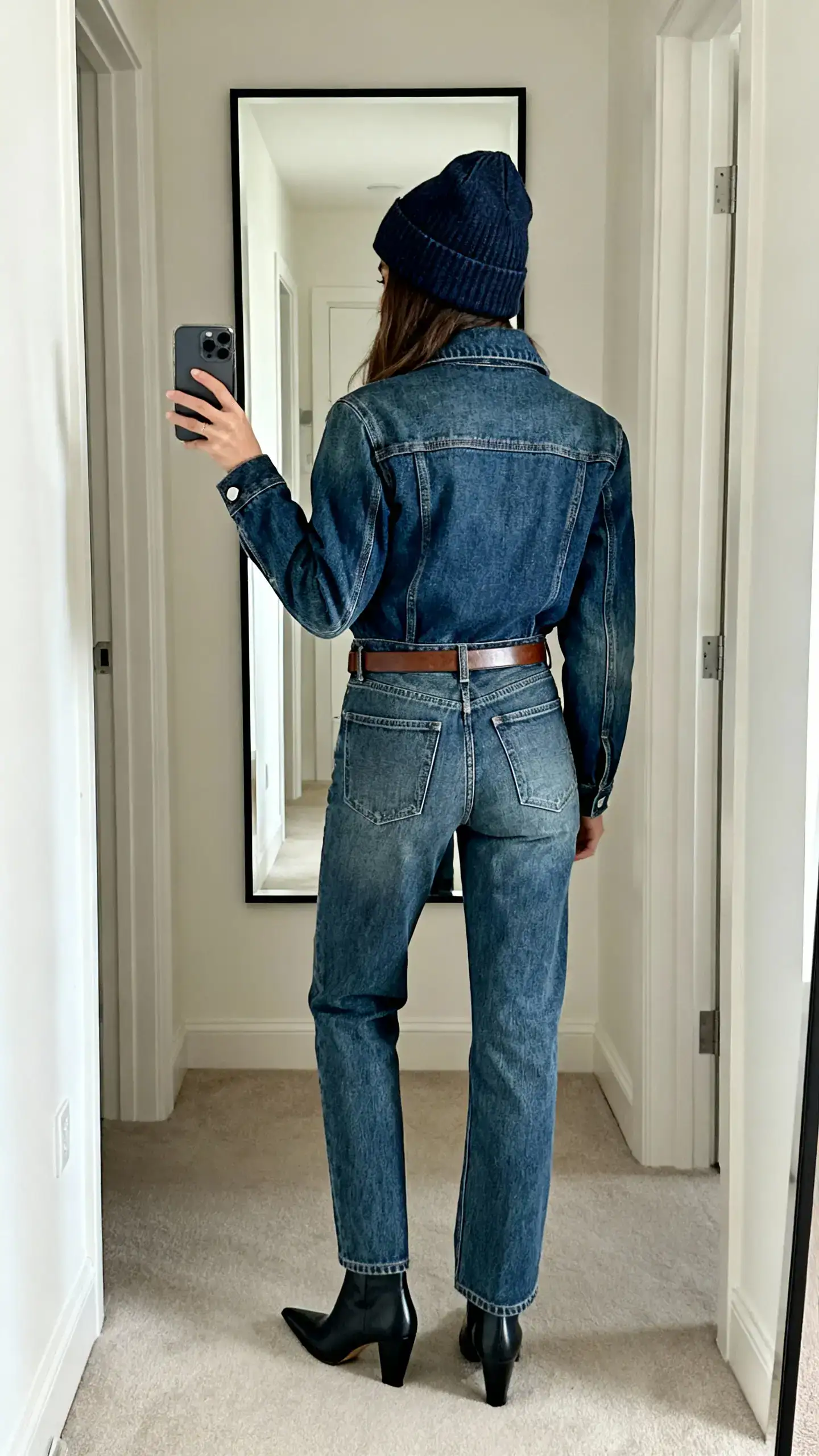 iPhone back view selfie of a woman wearing a dark denim beanie, structured indigo denim jacket, mid-wash straight jeans, pointed ankle boots, and a slim leather belt, face not visible, hallway mirror, soft indoor lighting, iPhone photo quality.