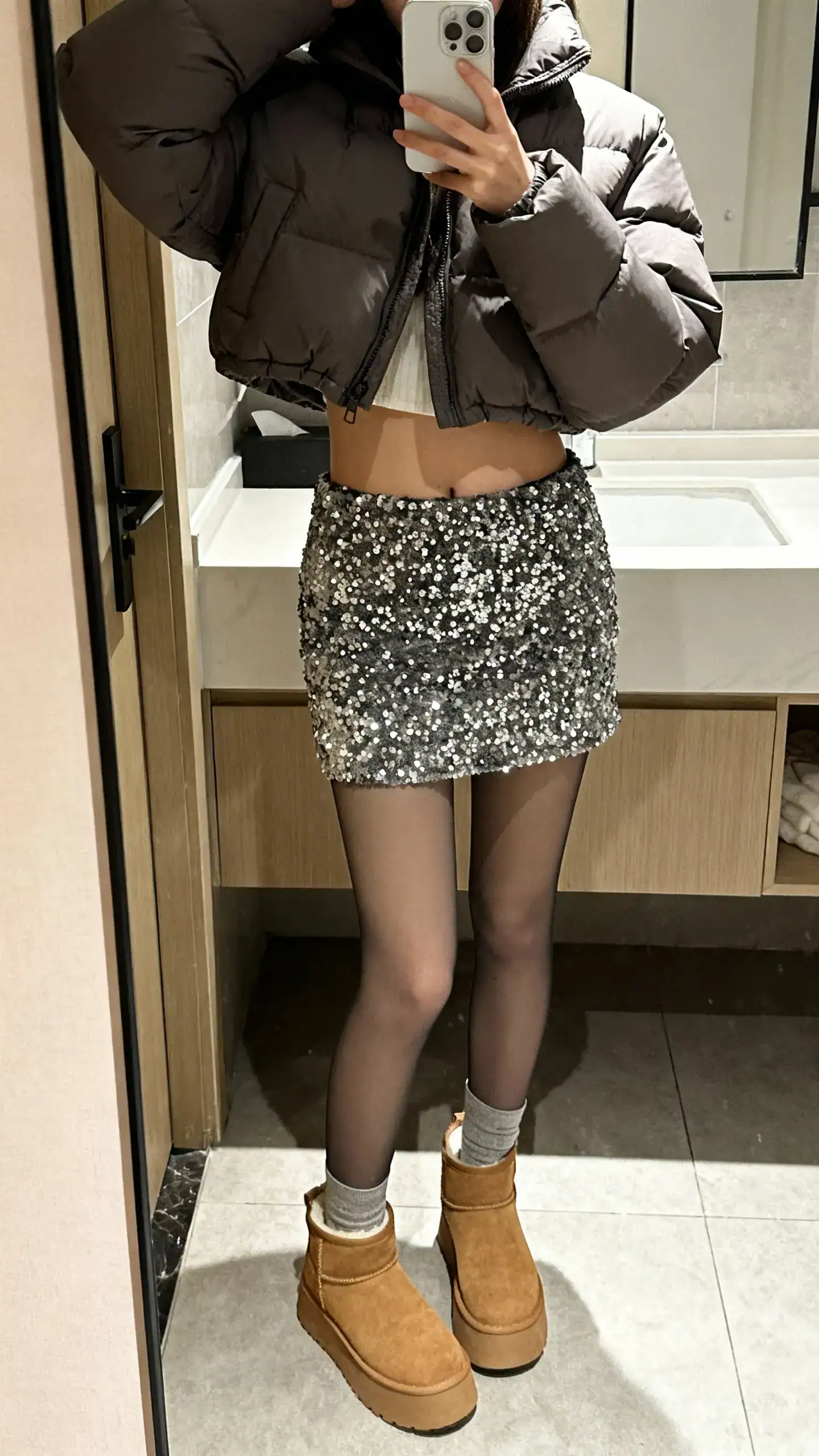 iPhone mirror selfie of a woman in a cropped puffer jacket with a sequin skirt, opaque tights, and chunky ankle boots, face not visible, bathroom mirror, soft indoor lighting, iPhone photo quality.