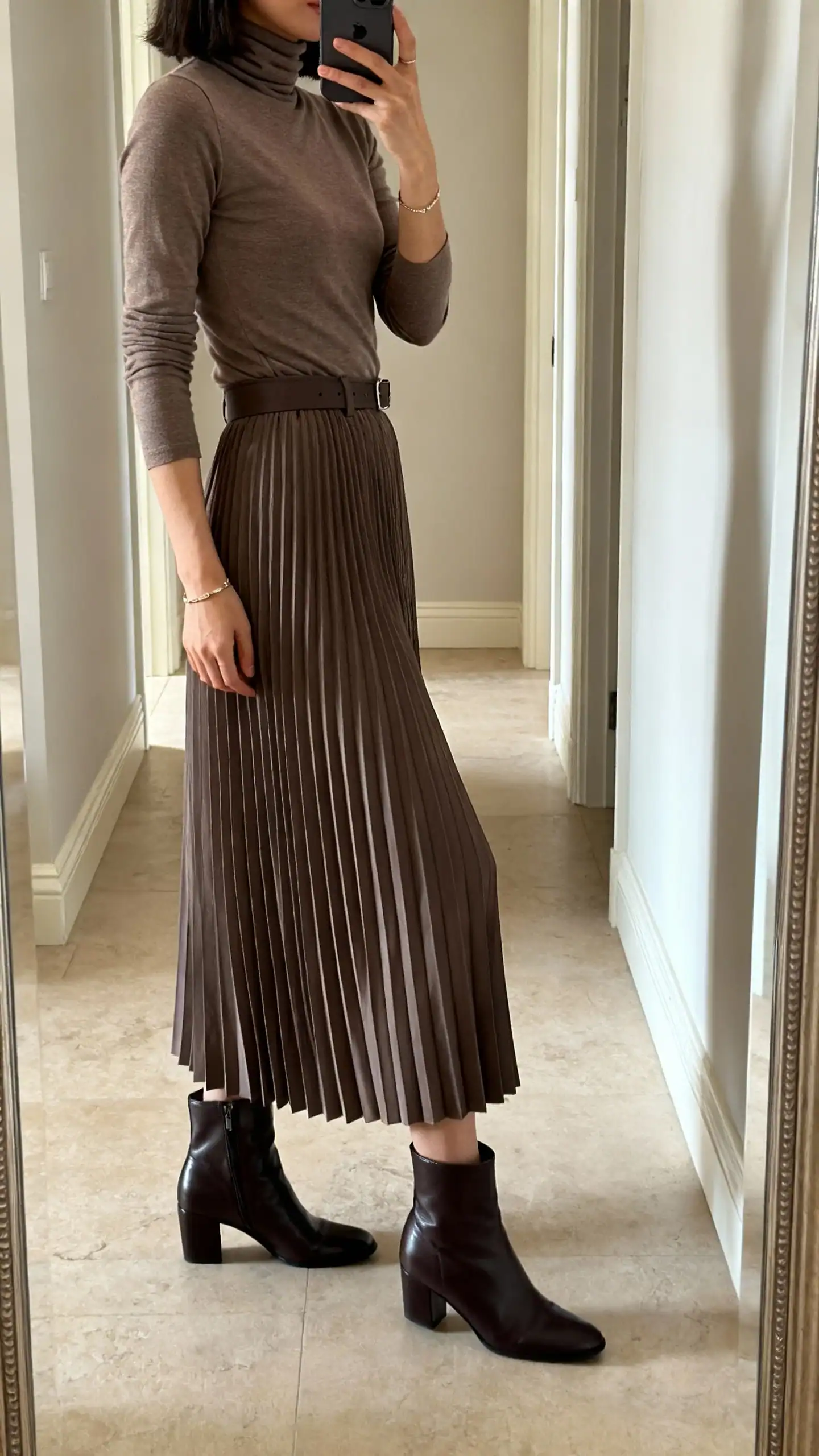 iPhone mirror selfie of a woman in a fitted turtleneck tucked into a pleated maxi skirt with sleek ankle boots and minimal jewelry, face not visible, side angle in a hallway full-length mirror, soft indoor lighting, iPhone photo quality.