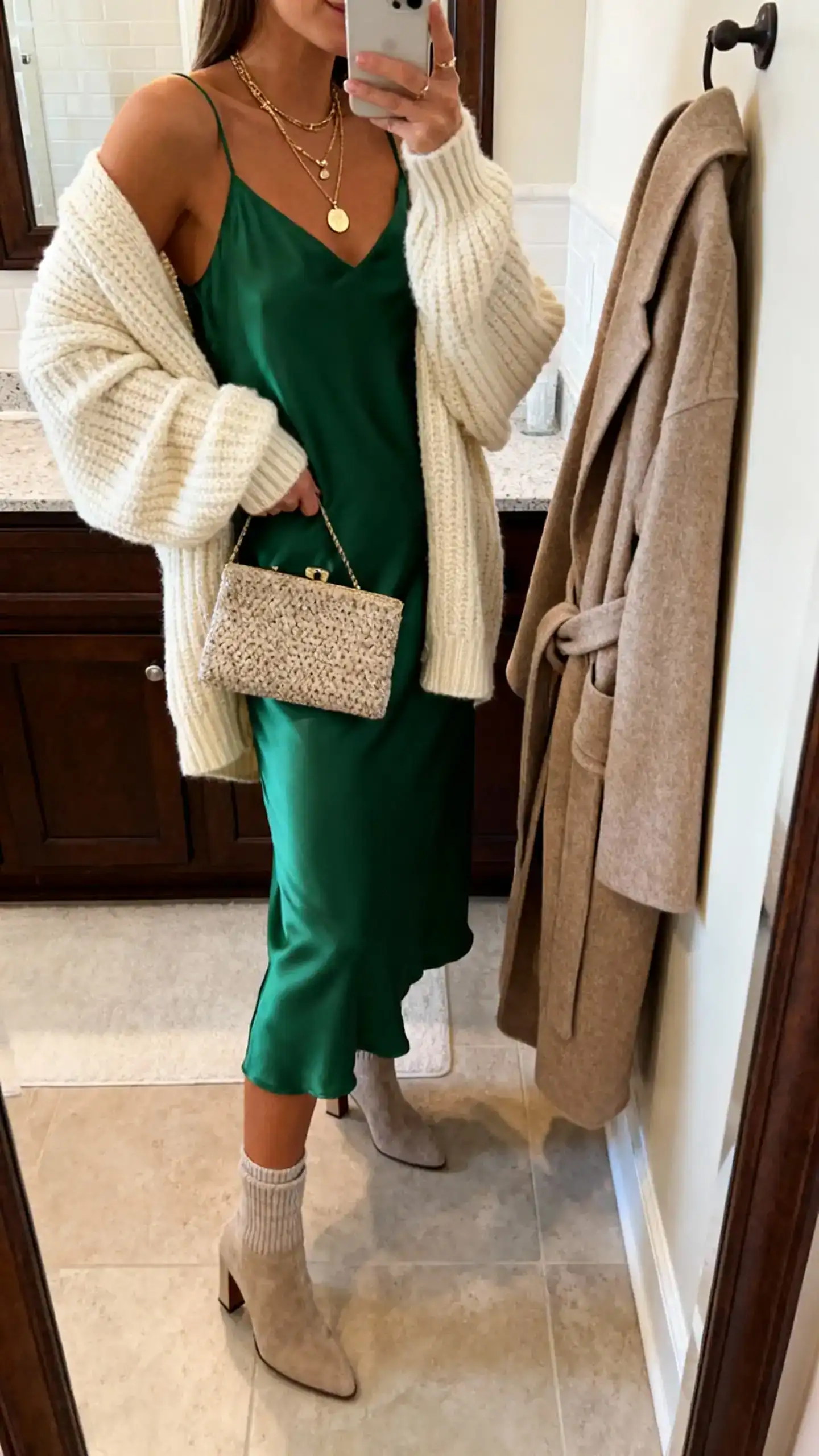 iPhone mirror selfie of a woman in a forest green satin midi slip dress with an oversized chunky cream cardigan, heeled sock boots, layered pendant necklaces, textured clutch, and a wool wrap coat over the hook; face not visible, side angle, bathroom mirror, warm indoor lighting, iPhone photo quality.