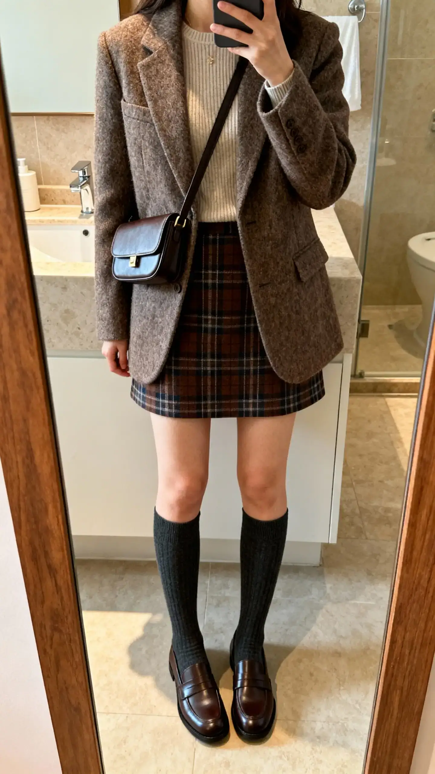 iPhone mirror selfie of a woman in a plaid mini skirt, wool blazer over a fine-knit top, knee-high socks and loafers with a structured crossbody, face not visible, bathroom mirror, warm indoor lighting, iPhone photo quality.