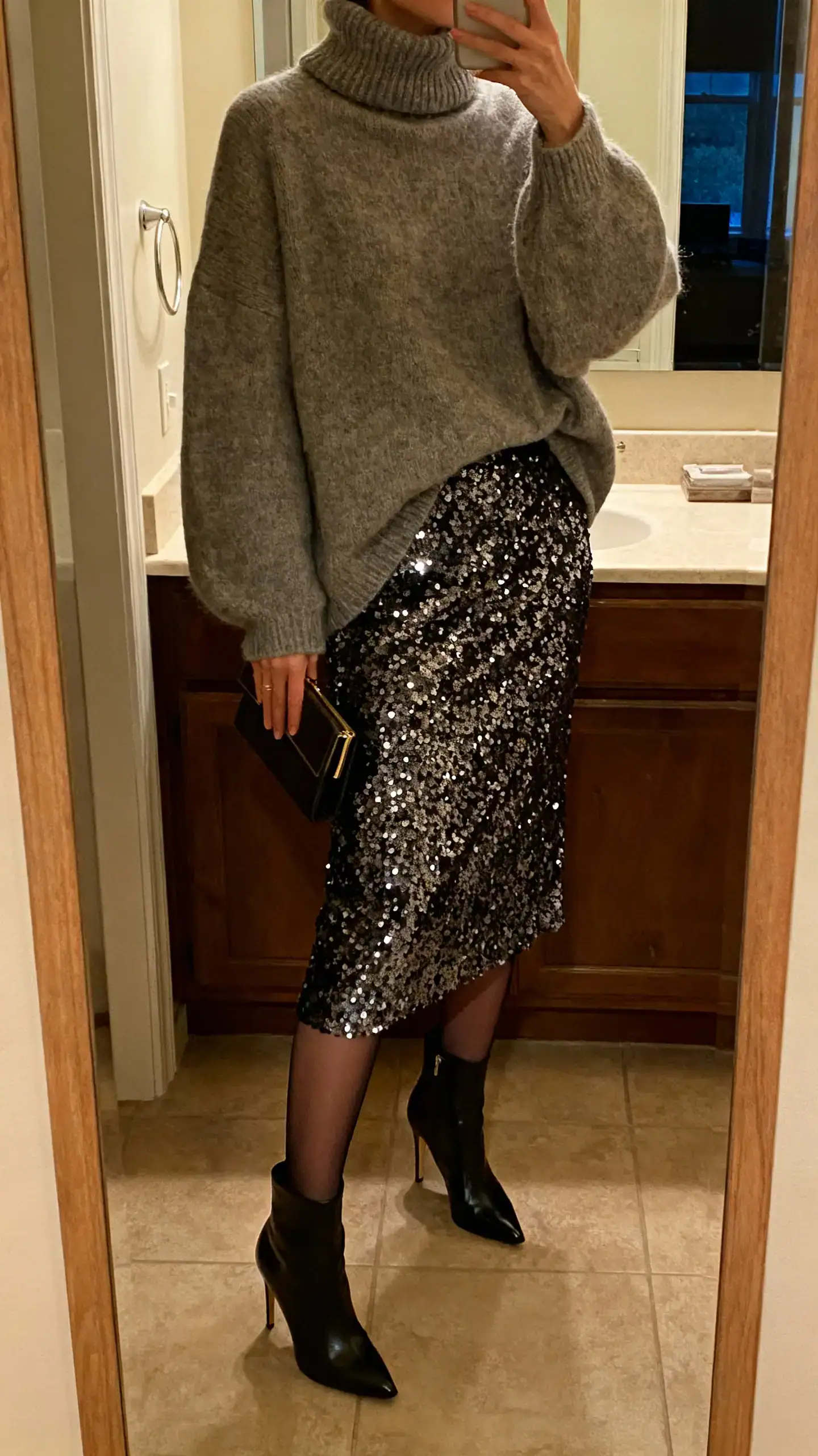 iPhone mirror selfie of a woman in a sequin midi skirt with an oversized turtleneck sweater, sheer tights, heeled boots and a small clutch, face not visible, cropped at neck, bathroom mirror, warm evening indoor lighting, iPhone photo quality.