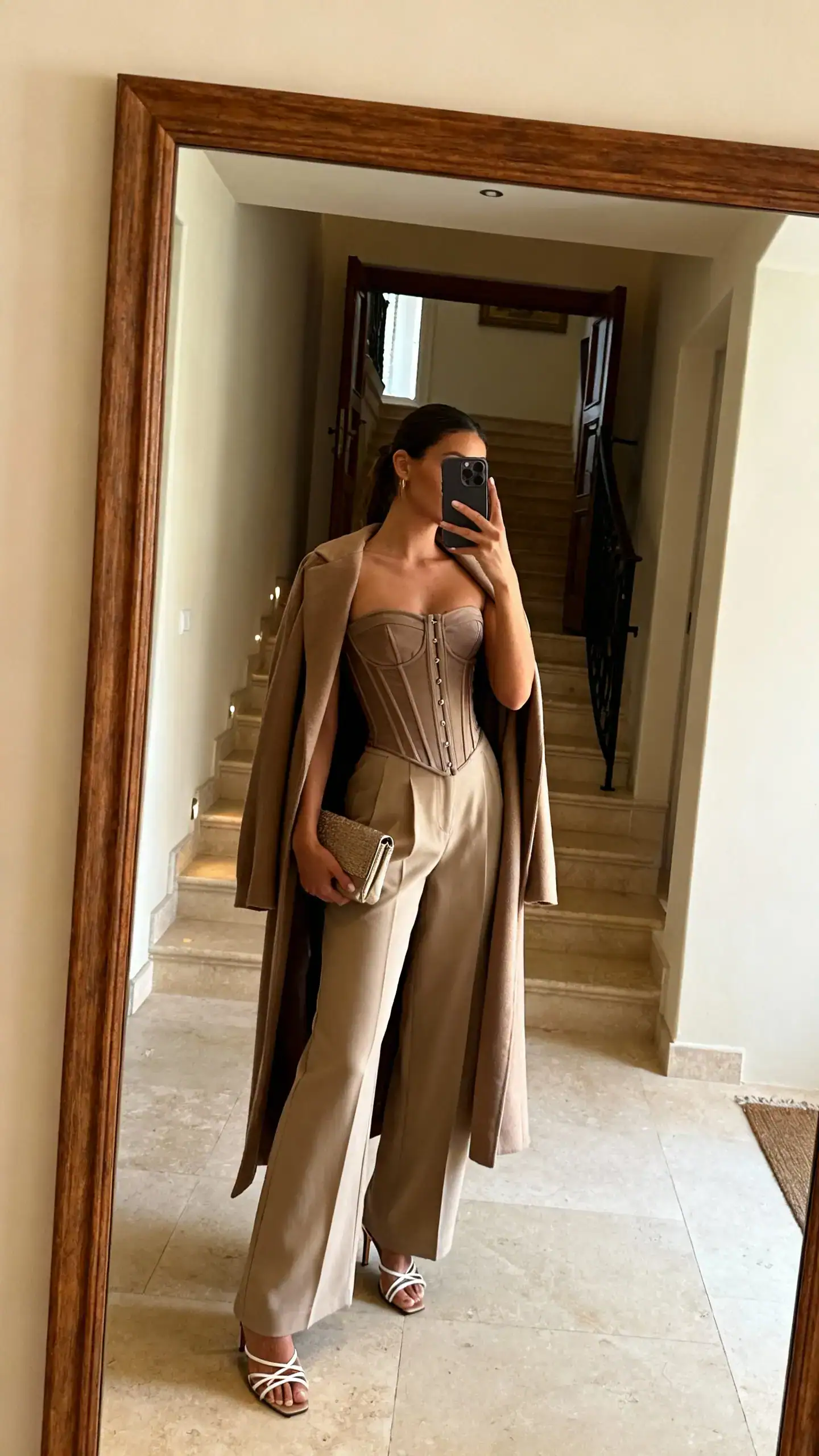 iPhone mirror selfie of a woman in a structured corset top with wide-leg pants and a longline coat draped over shoulders, strappy heels and small clutch, face not visible, side angle, foyer mirror, warm indoor lighting, iPhone photo quality.