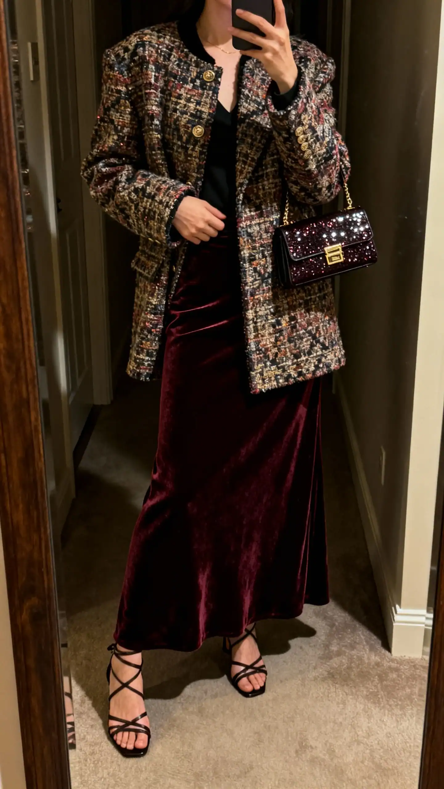 iPhone mirror selfie of a woman in a velvet maxi skirt with a statement coat, strappy heels, and a small evening bag, face not visible, cropped at neck in a dim hallway mirror, warm indoor lighting, iPhone photo quality.
