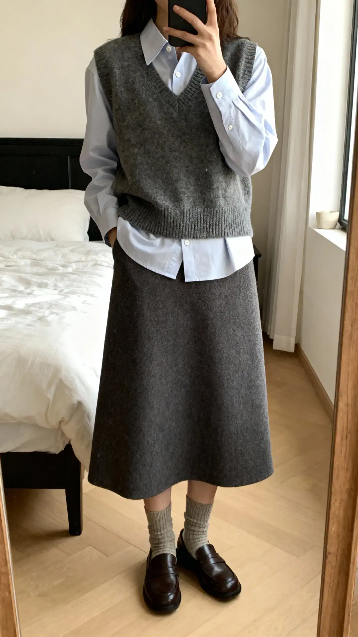 iPhone mirror selfie of a woman in a wool A-line skirt with a crisp button-down layered under a sweater vest, loafers and crew socks, face not visible, cropped at shoulders, bedroom mirror, soft natural light, iPhone photo quality.