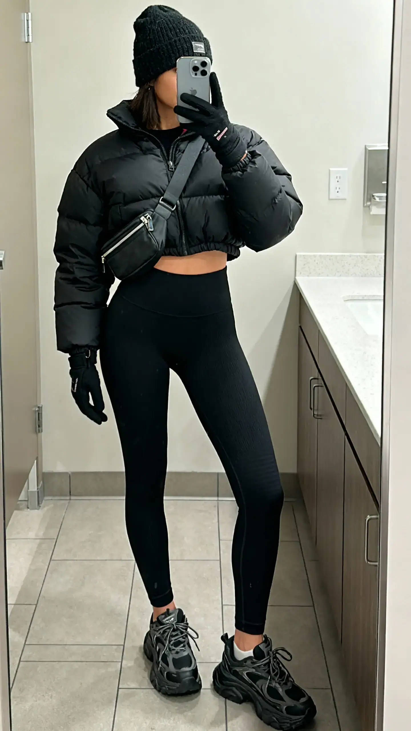iPhone mirror selfie of a woman in black fleece-lined thermal leggings, a black cropped puffer jacket, chunky sneakers, a cozy beanie, touchscreen gloves, and a crossbody belt bag worn across the chest, face not visible, bathroom mirror, indoor lighting, iPhone photo quality.