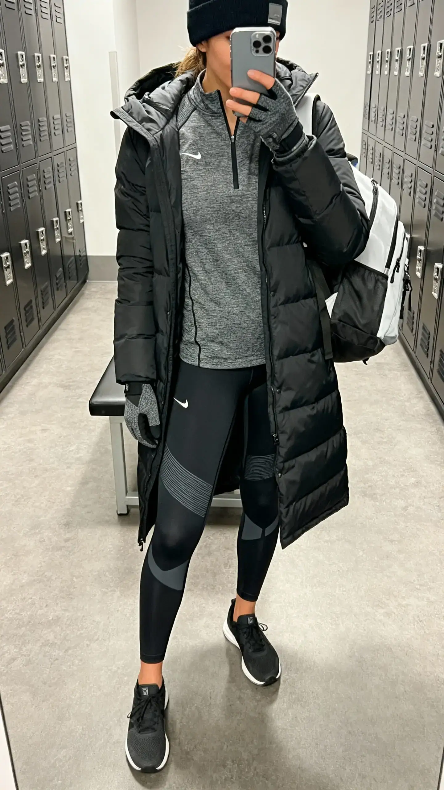 iPhone mirror selfie of a woman in compression performance leggings, a half-zip active top, a streamlined mid-thigh parka, minimal trainers, a sporty backpack, and tech-friendly gloves with a beanie, face not visible, cropped at shoulders, gym locker room mirror, indoor lighting, iPhone photo quality.
