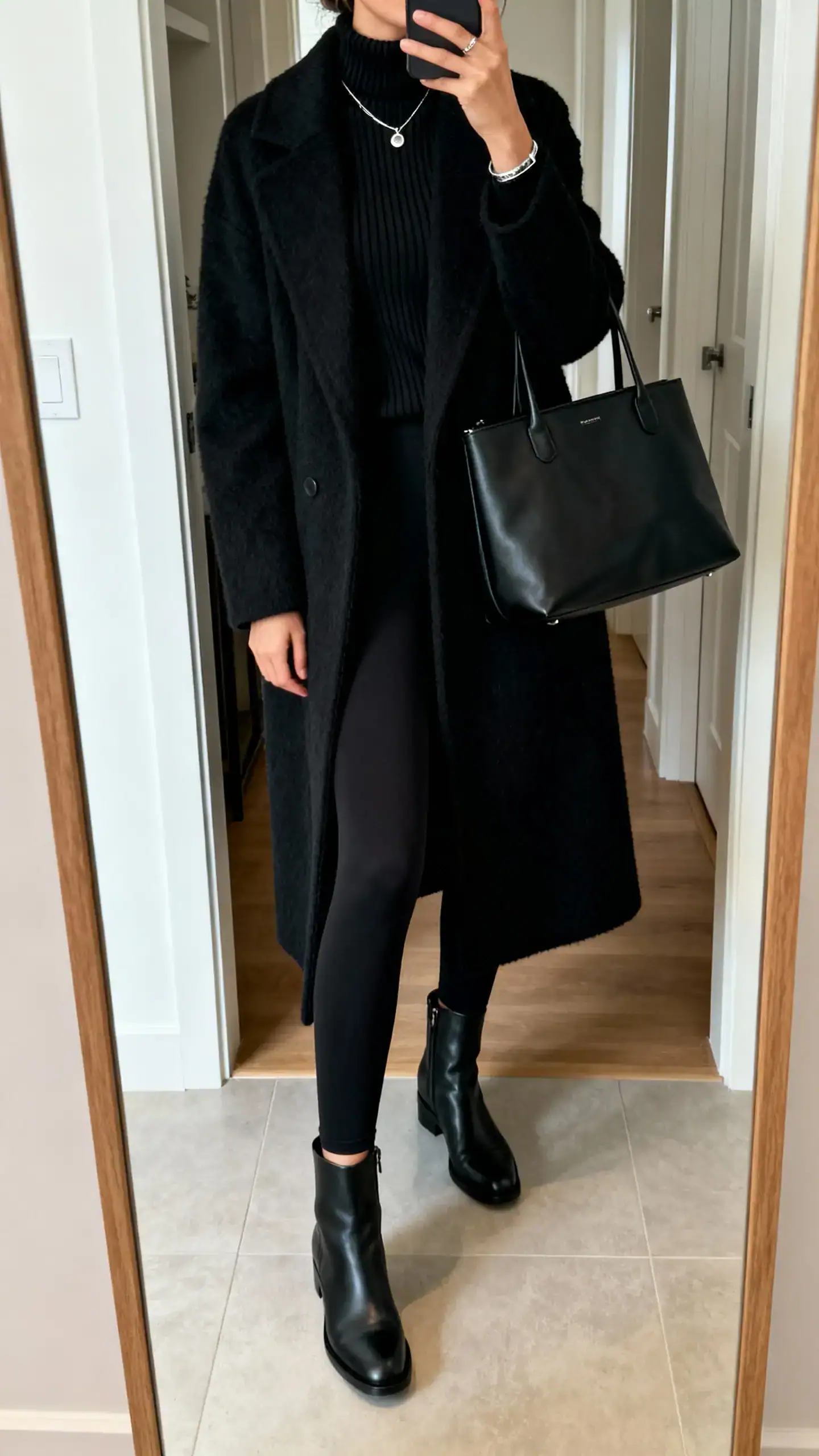 iPhone mirror selfie of a woman in head-to-toe black: opaque black leggings, black ribbed turtleneck, black ankle boots, a long black wool coat, a matte black tote, and subtle silver jewelry, face not visible, cropped at neck, entryway full-length mirror, natural indoor light, iPhone photo quality.