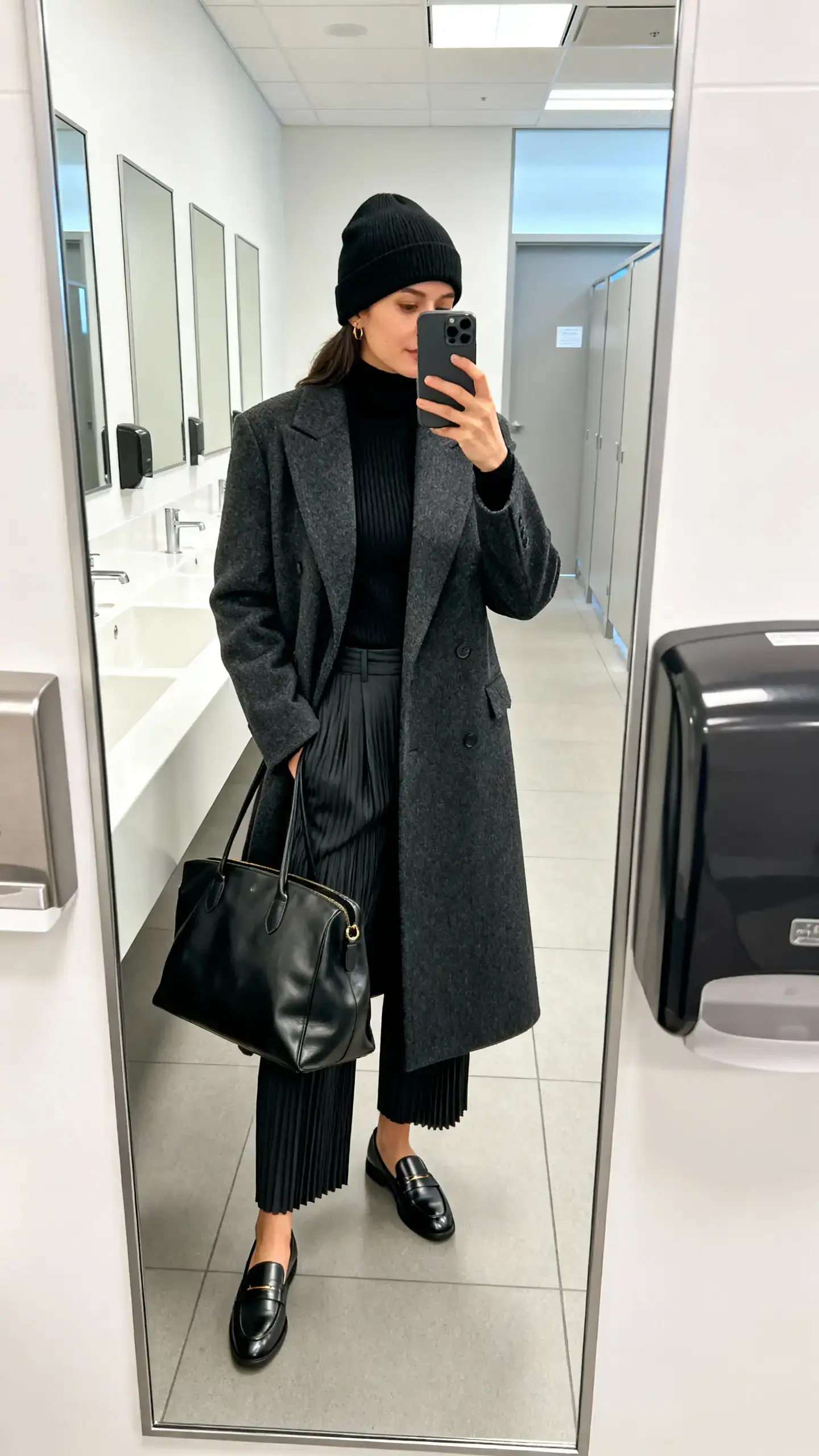 iPhone mirror selfie of a woman wearing a black fitted beanie, tailored charcoal wool coat, fine-gauge black turtleneck, high-waist pleated trousers, sleek loafers, and a leather tote, face not visible, cropped at neck, office bathroom mirror, bright indoor lighting, iPhone photo quality.