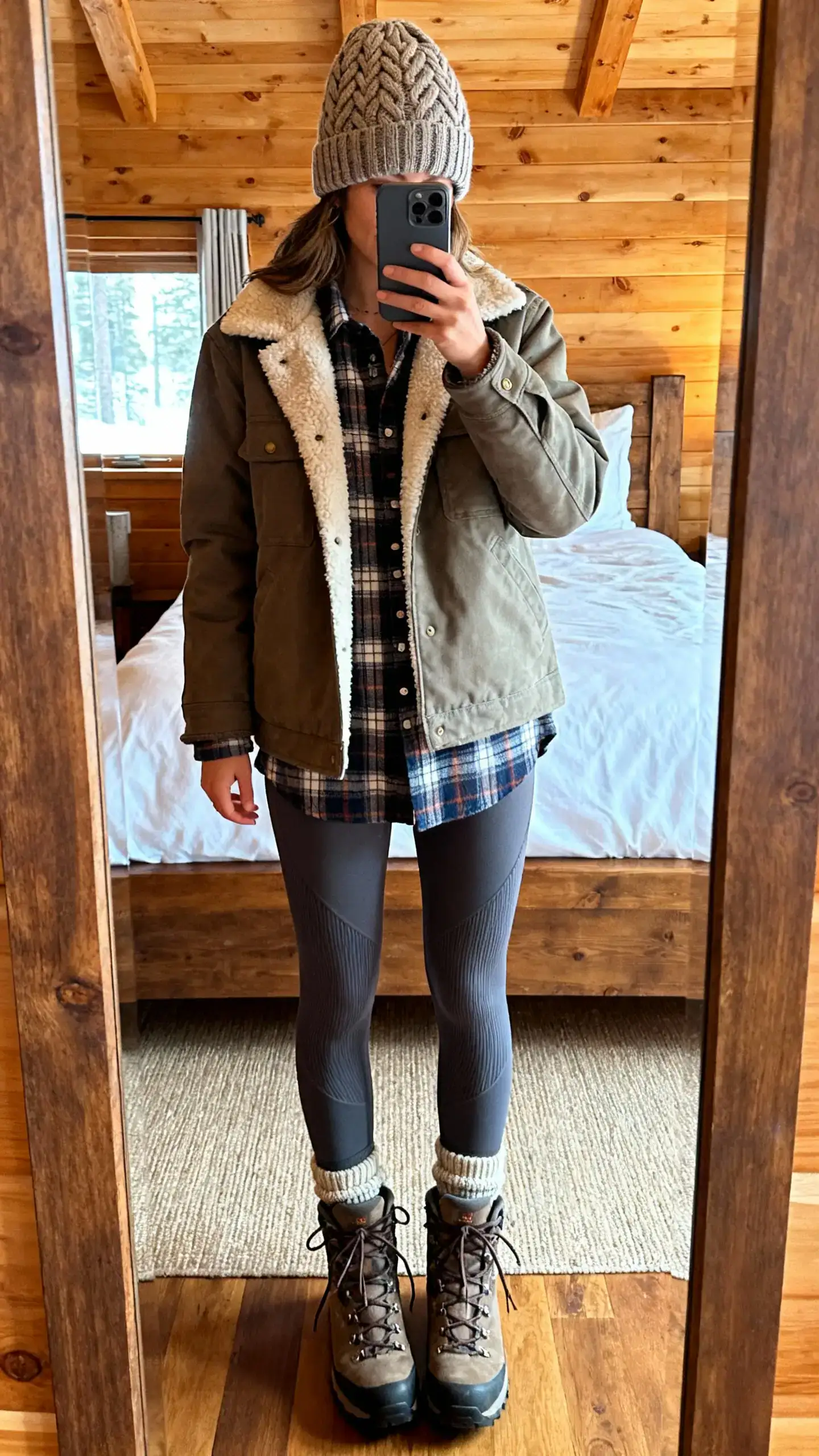 iPhone mirror selfie of a woman wearing a cable-knit beanie, sherpa-lined chore jacket, plaid flannel shirt, thermal leggings, lace-up hiking boots, and wool socks peeking out, face not visible, cropped at shoulders, cabin-style bedroom mirror, warm indoor light, iPhone photo quality.