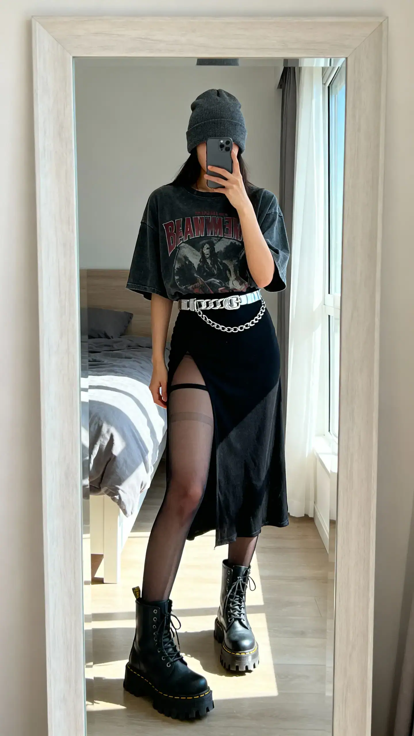 iPhone mirror selfie of a woman wearing a charcoal beanie, oversized band tee, black slip dress layered over sheer tights, lug-sole combat boots, and a silver chain belt, face not visible, bedroom mirror, afternoon window light, iPhone photo quality.