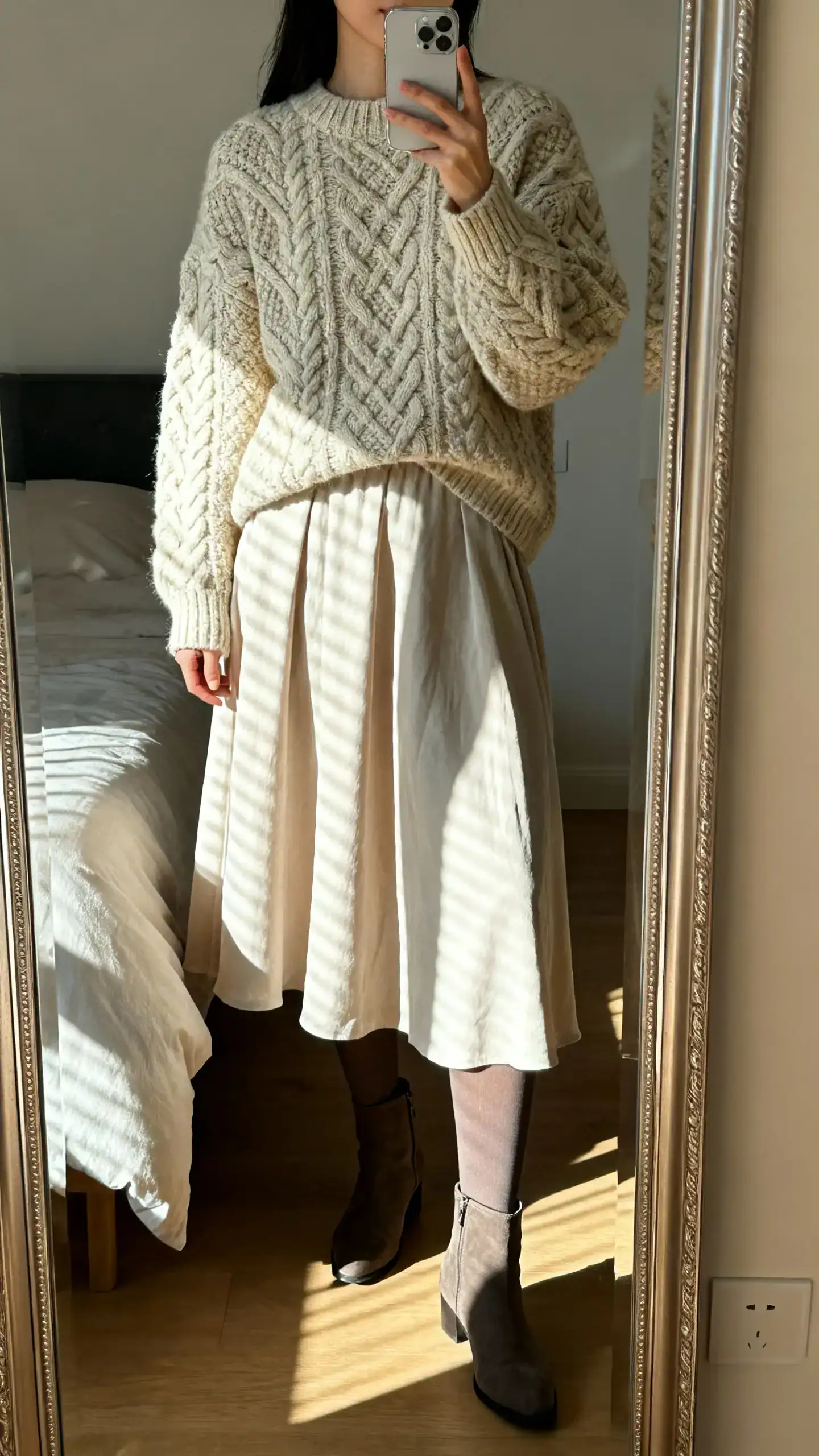 iPhone mirror selfie of a woman wearing a chunky cable-knit sweater and a flowy midi skirt with opaque tights and ankle boots, face not visible, cropped at neck, bedroom mirror with morning window light, iPhone photo quality.