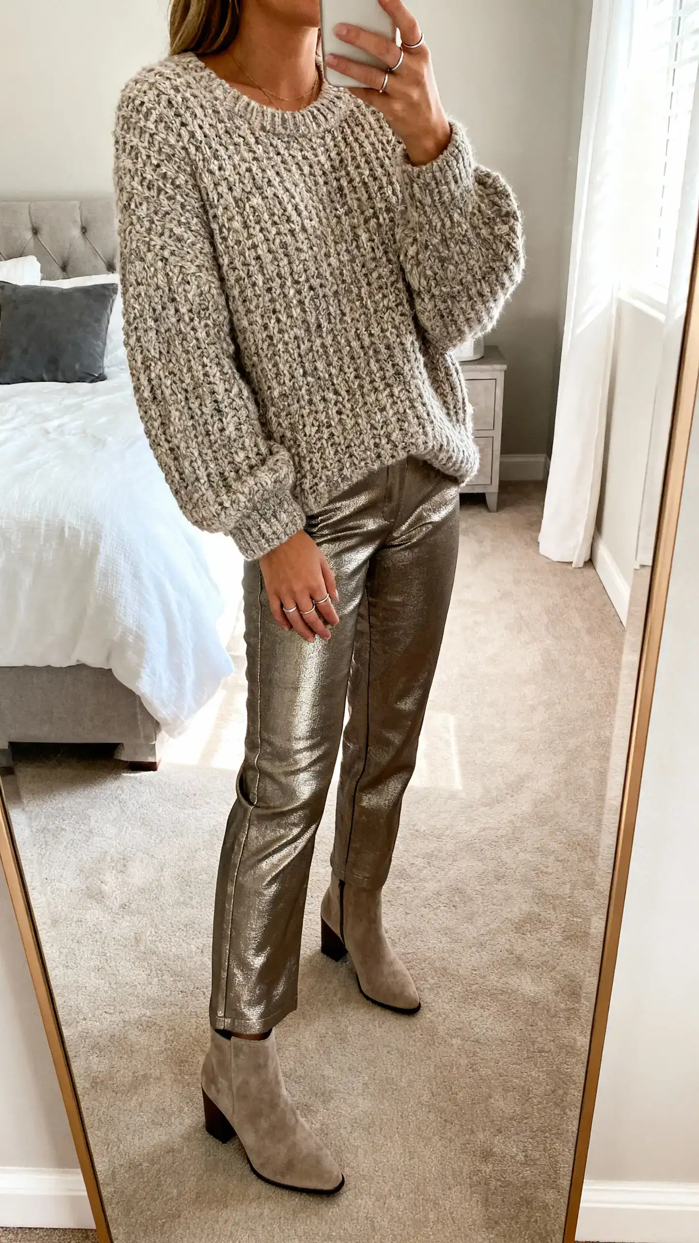 iPhone mirror selfie of a woman wearing a chunky textured sweater with metallic pants, block-heel boots, and simple rings, face not visible, cropped at neck, bedroom mirror, natural daylight, iPhone photo quality.