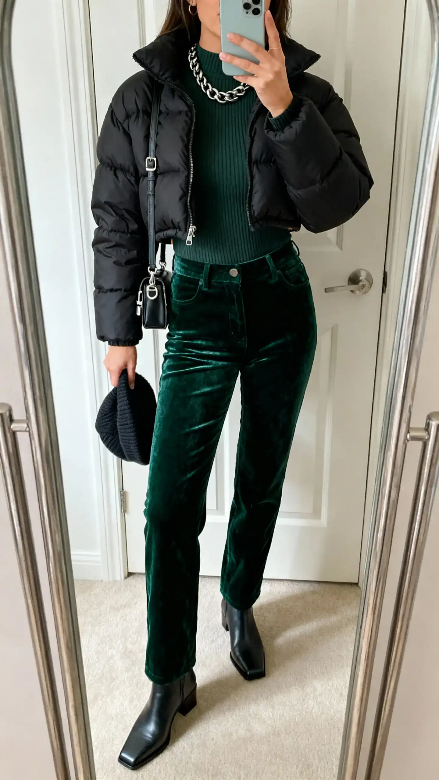 iPhone mirror selfie of a woman wearing a cropped black puffer jacket, deep green velvet straight-leg pants, fitted knit bodysuit, square-toe boots, chunky chain necklace, small shoulder bag with metal hardware, and a beanie in hand; face not visible, full-length mirror by the door, natural daylight, iPhone photo quality.