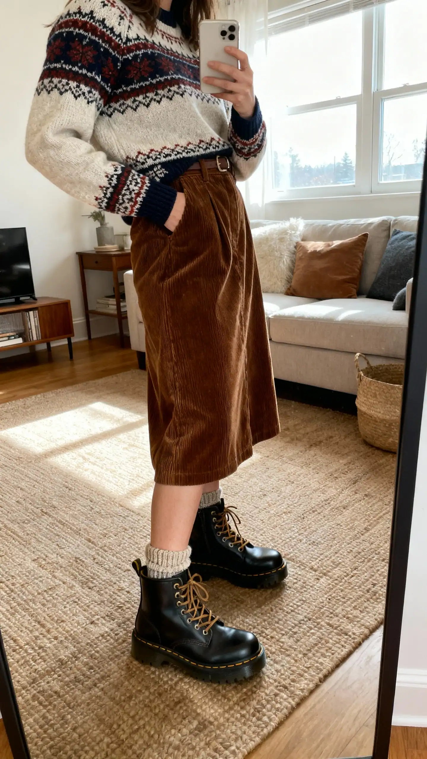 iPhone mirror selfie of a woman wearing a Fair Isle sweater tucked into a corduroy midi skirt with chunky lace-up boots and wool socks peeking out, face not visible, side angle in a cozy living room mirror, natural window light, iPhone photo quality.