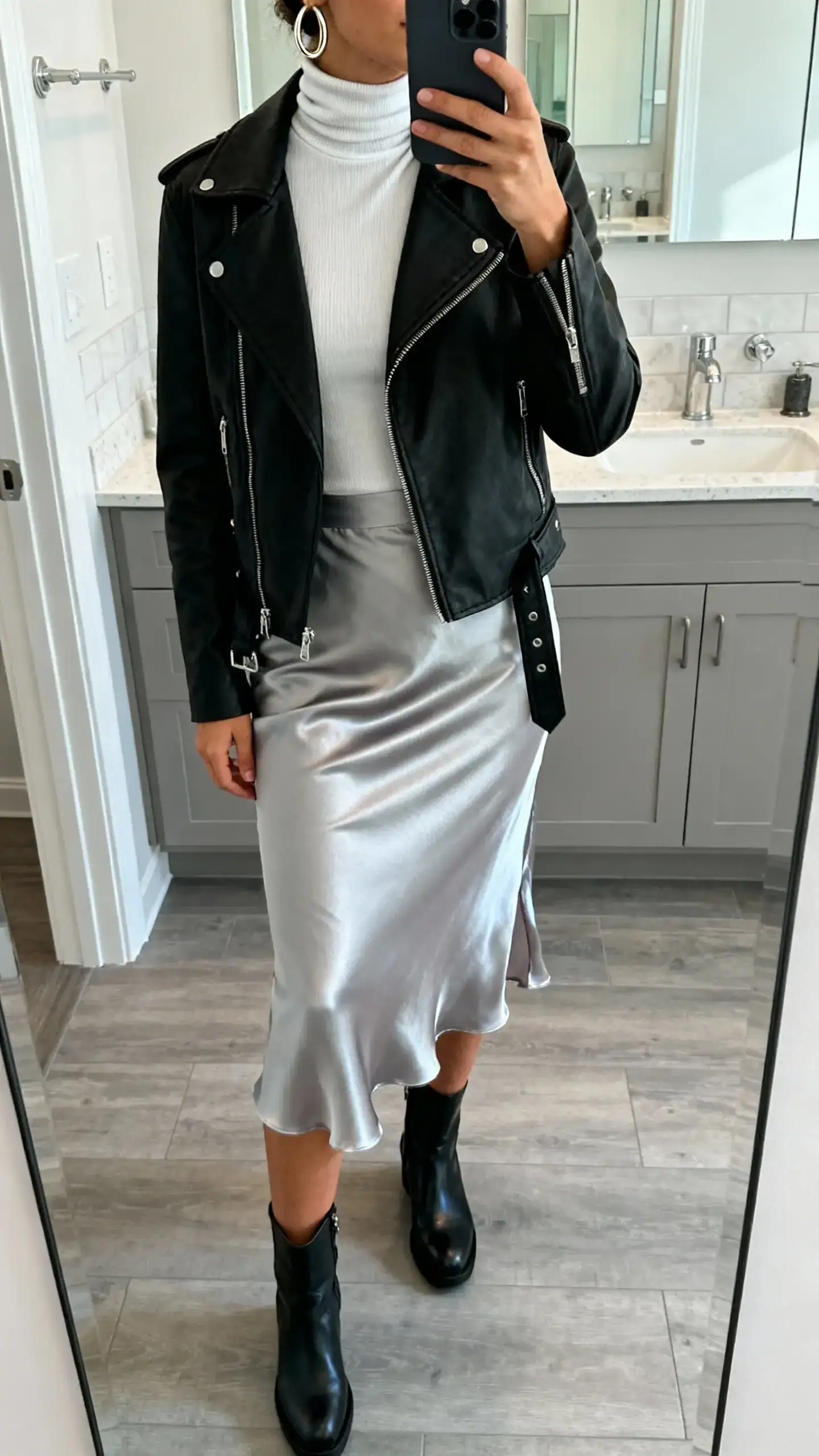 iPhone mirror selfie of a woman wearing a fitted turtleneck tucked into a satin slip skirt with a black moto jacket, ankle boots, and hoop earrings, face not visible, cropped at neck, bathroom mirror, cool indoor lighting, iPhone photo quality.