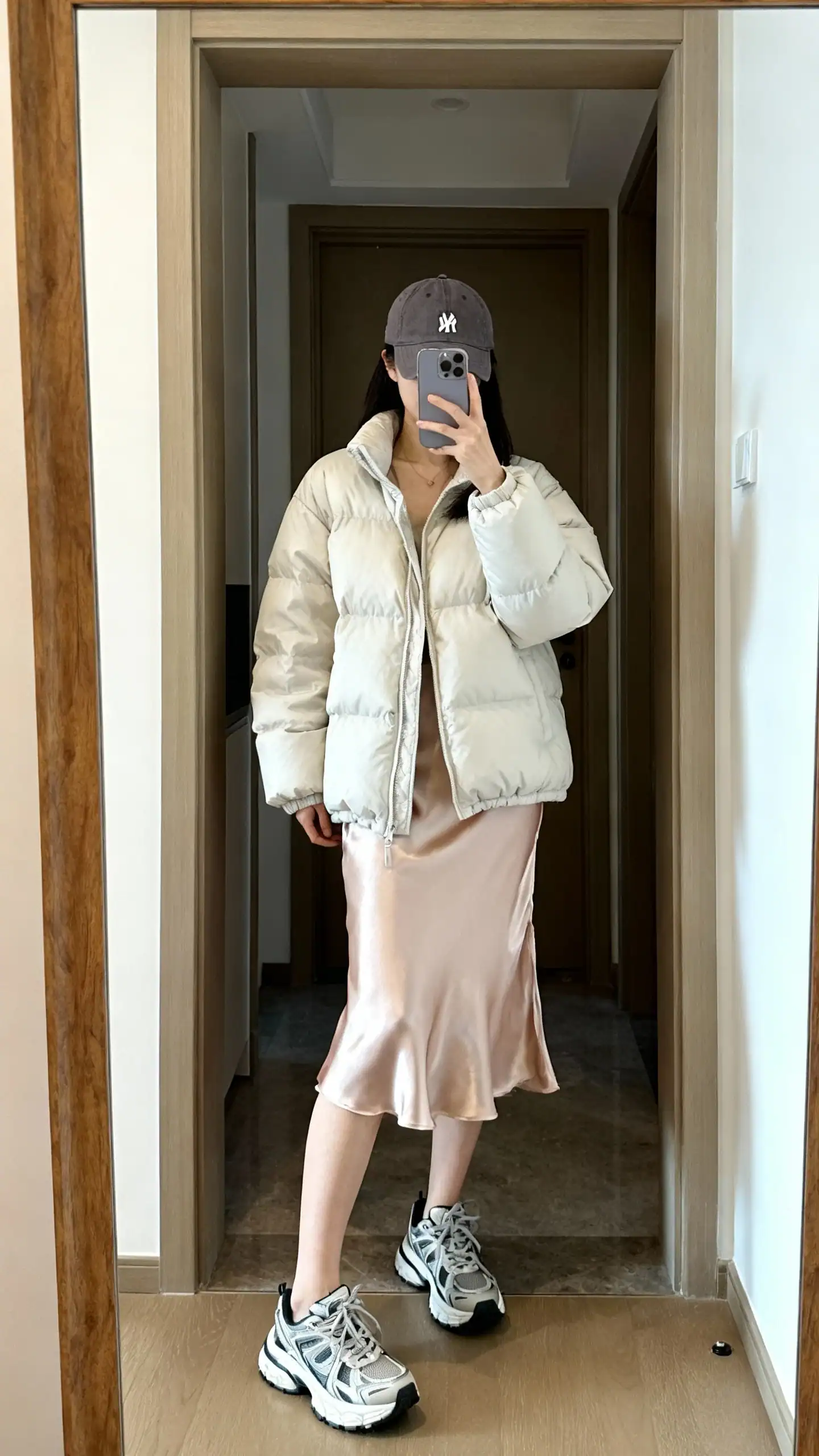 iPhone mirror selfie of a woman wearing a lightweight puffer jacket over a silk slip skirt with chunky sneakers and a baseball cap, face not visible, cropped at neck in an entryway mirror, natural indoor light, iPhone photo quality.