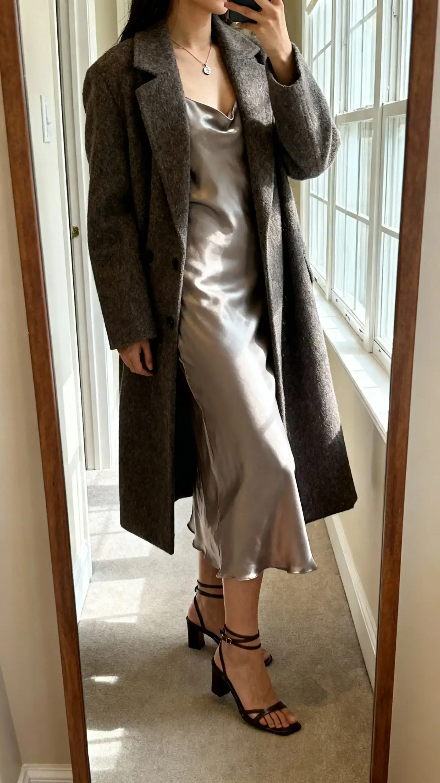 iPhone mirror selfie of a woman wearing a long wool coat layered over a satin slip dress with delicate pendant necklace and ankle-strap heels, face not visible, side angle in hallway mirror, morning window light, iPhone photo quality.