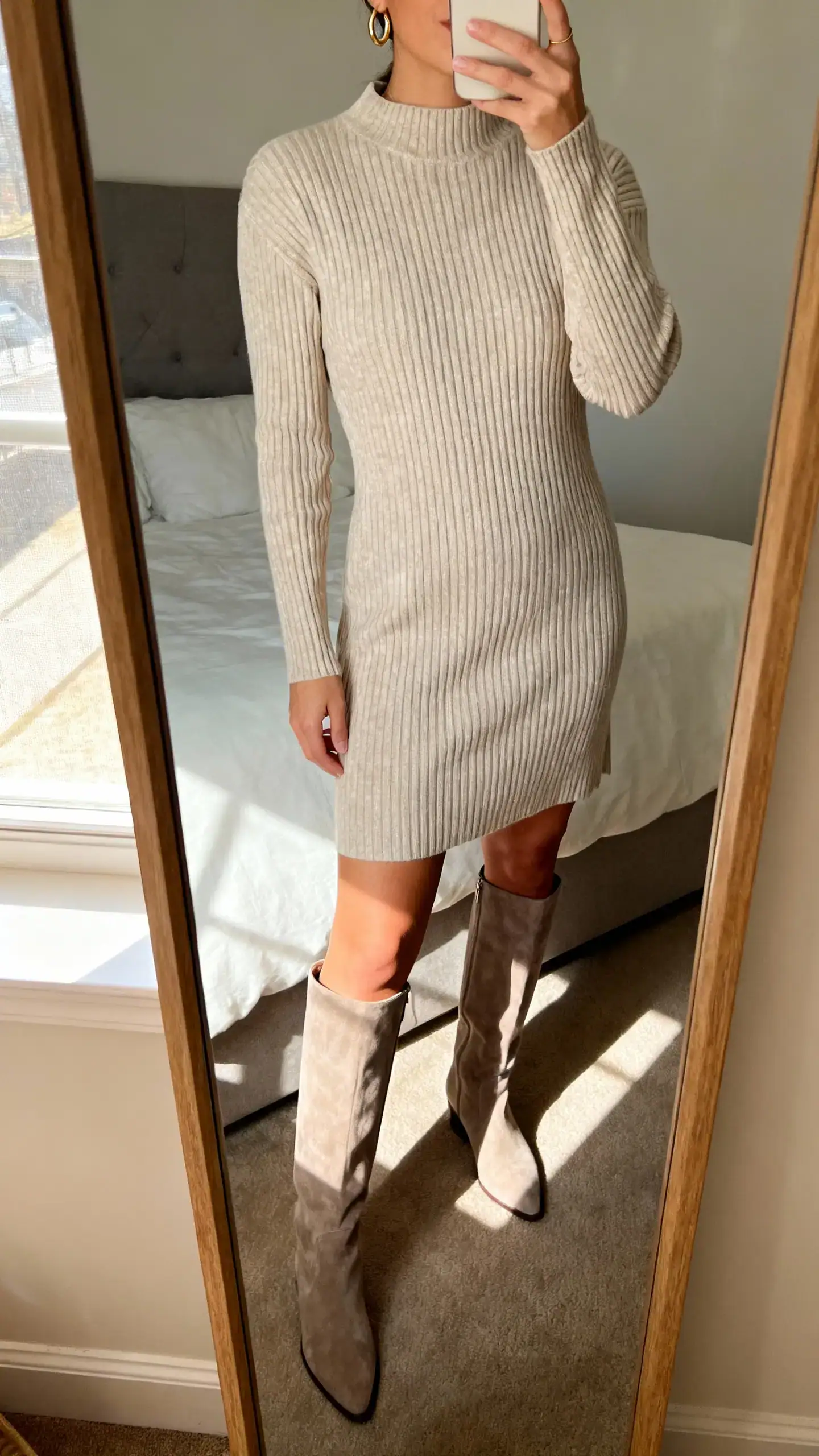 iPhone mirror selfie of a woman wearing a luxe ribbed knit dress with knee-high boots and minimal gold hoops, face not visible, cropped at neck, bedroom mirror, soft morning window light, iPhone photo quality.