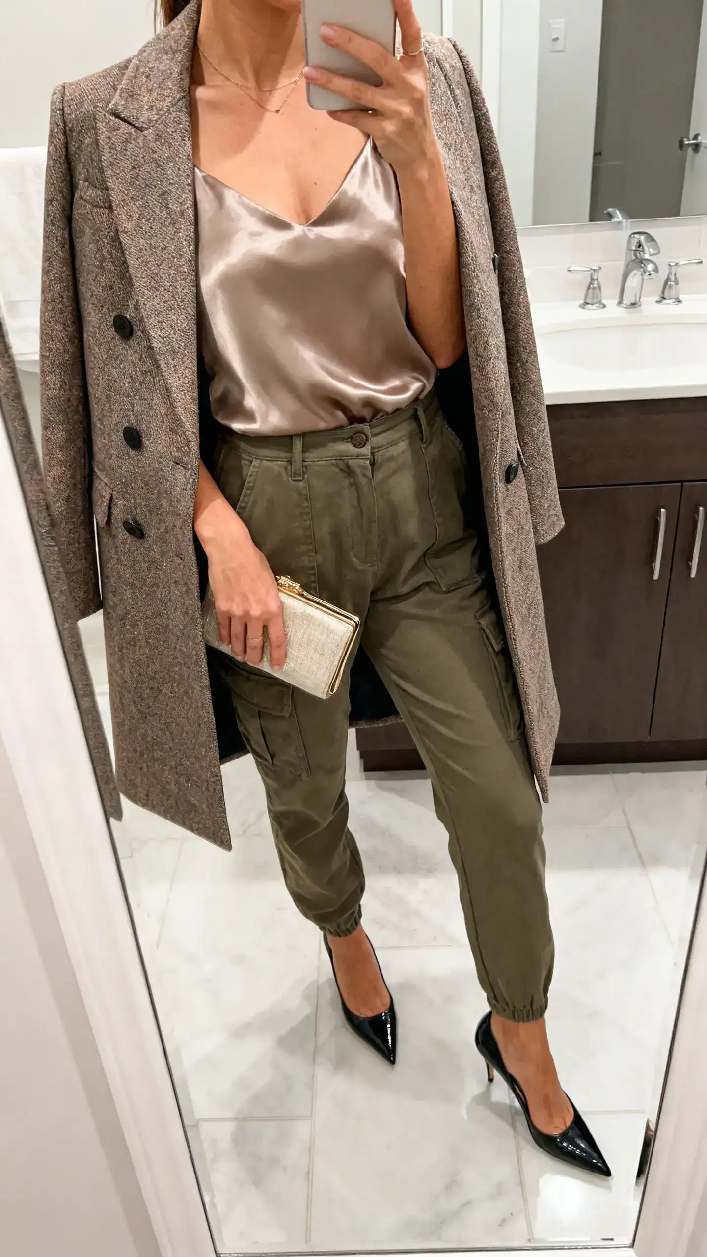 iPhone mirror selfie of a woman wearing a satin top tucked into cargo pants with a dressy tailored coat, pointed-toe heels, and a clutch, face not visible, cropped at neck, bathroom mirror, bright indoor lighting, iPhone photo quality.