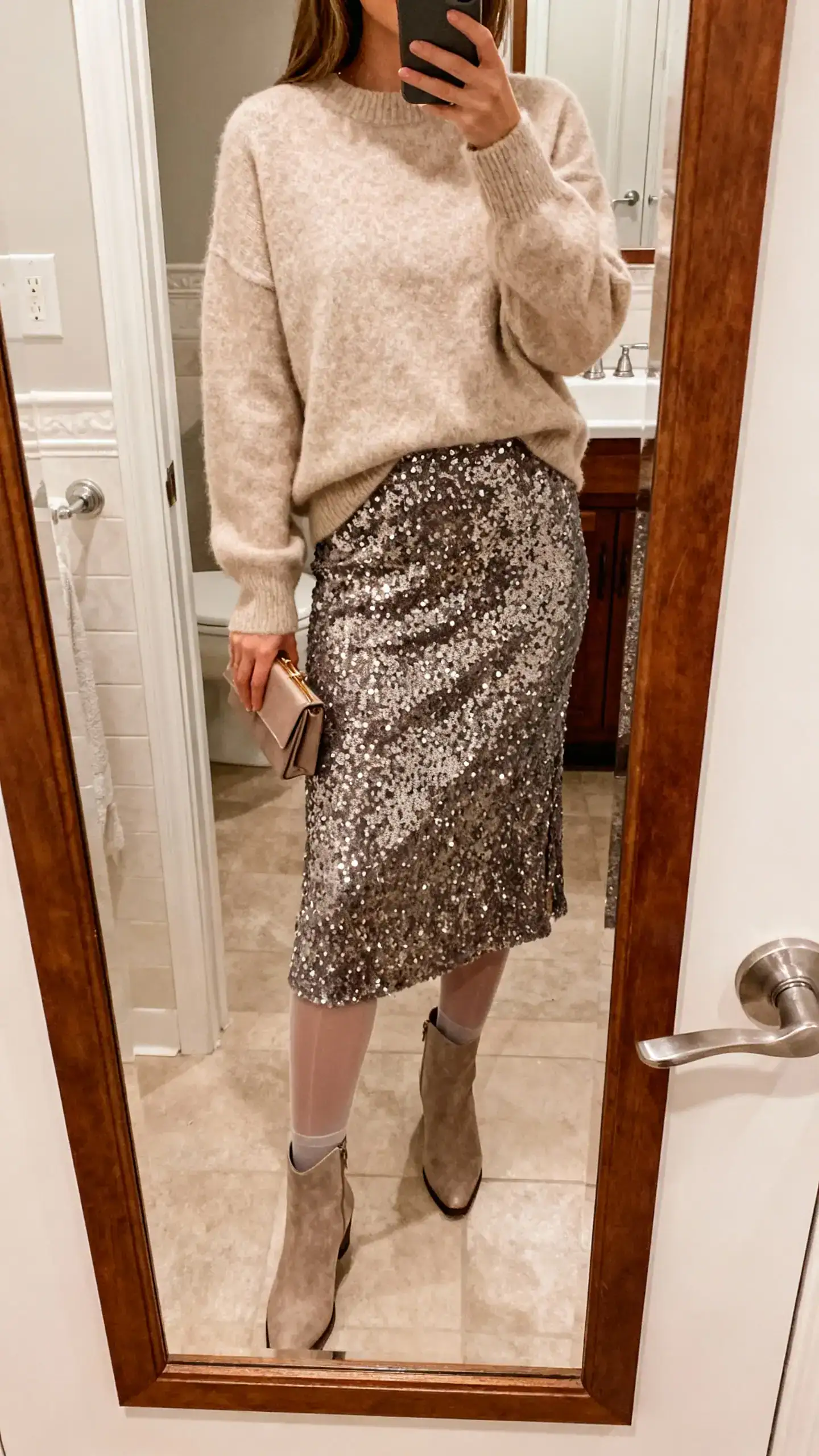 iPhone mirror selfie of a woman wearing a sequin midi skirt with a plush cashmere crewneck sweater, sheer tights, ankle boots, and a small clutch, face not visible, bathroom mirror, warm indoor lighting, iPhone photo quality.