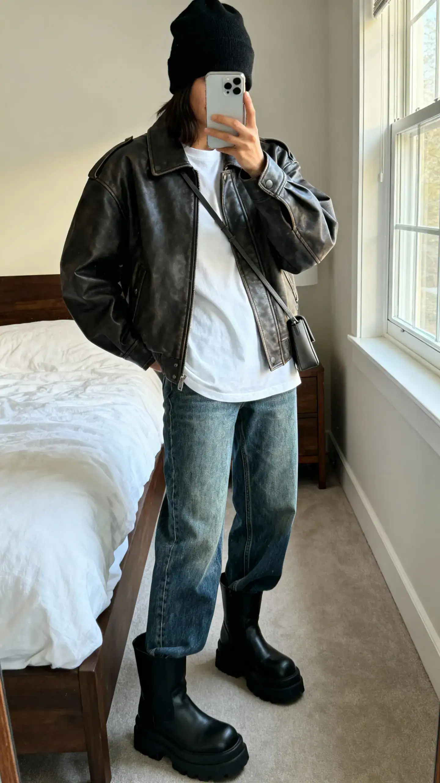 iPhone mirror selfie of a woman wearing a slouchy black beanie, oversized leather moto jacket, plain white tee, straight-leg vintage blue jeans, chunky black boots, and a minimalist crossbody, face not visible, cropped at neck, bedroom mirror, morning window light, iPhone photo quality.