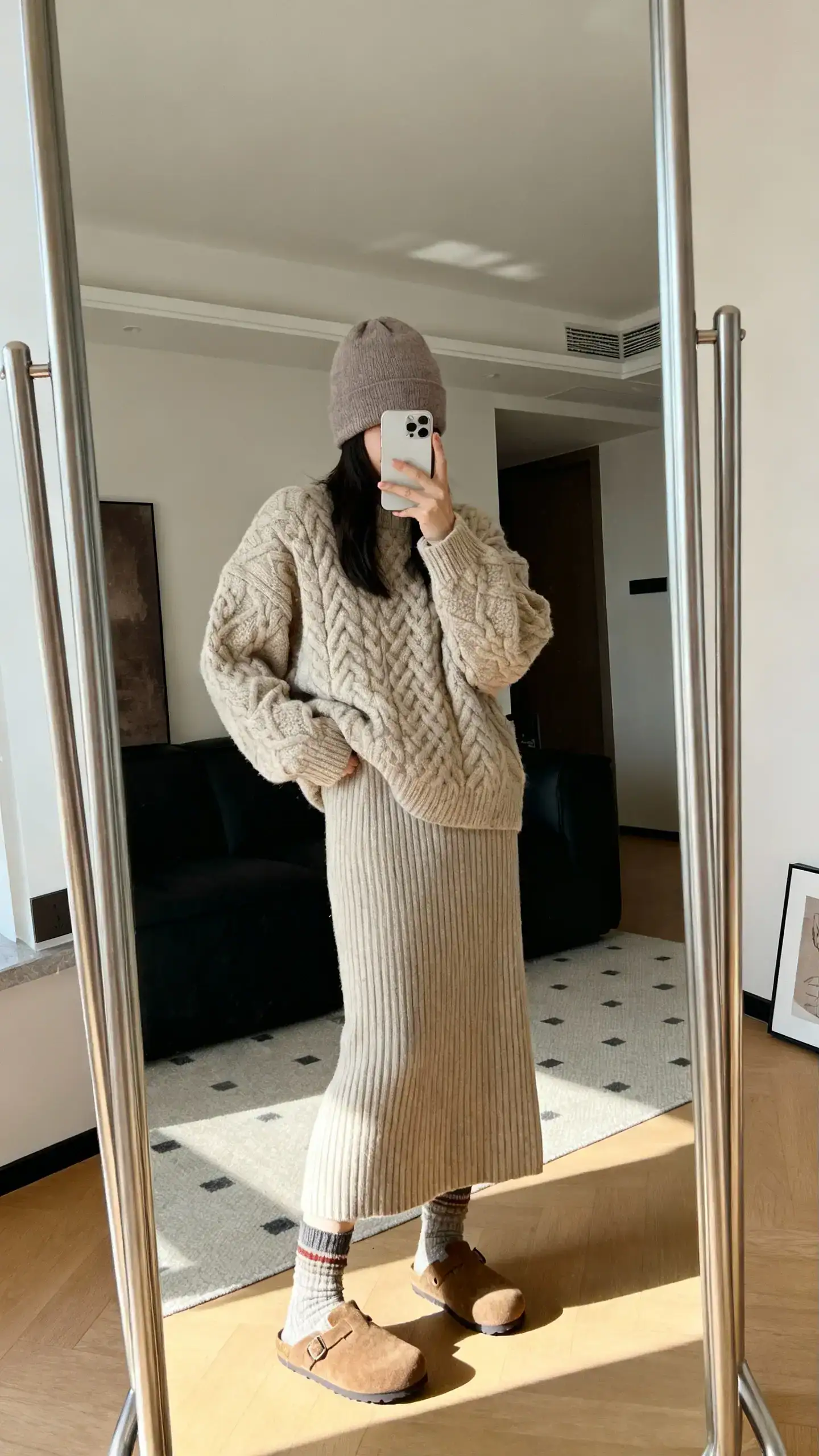 iPhone mirror selfie of a woman wearing a soft taupe beanie, chunky oatmeal cable-knit sweater, matching ribbed knit skirt, cozy crew socks, and suede clogs, face not visible, cropped at shoulders, living room full-length mirror, gentle morning light, iPhone photo quality.