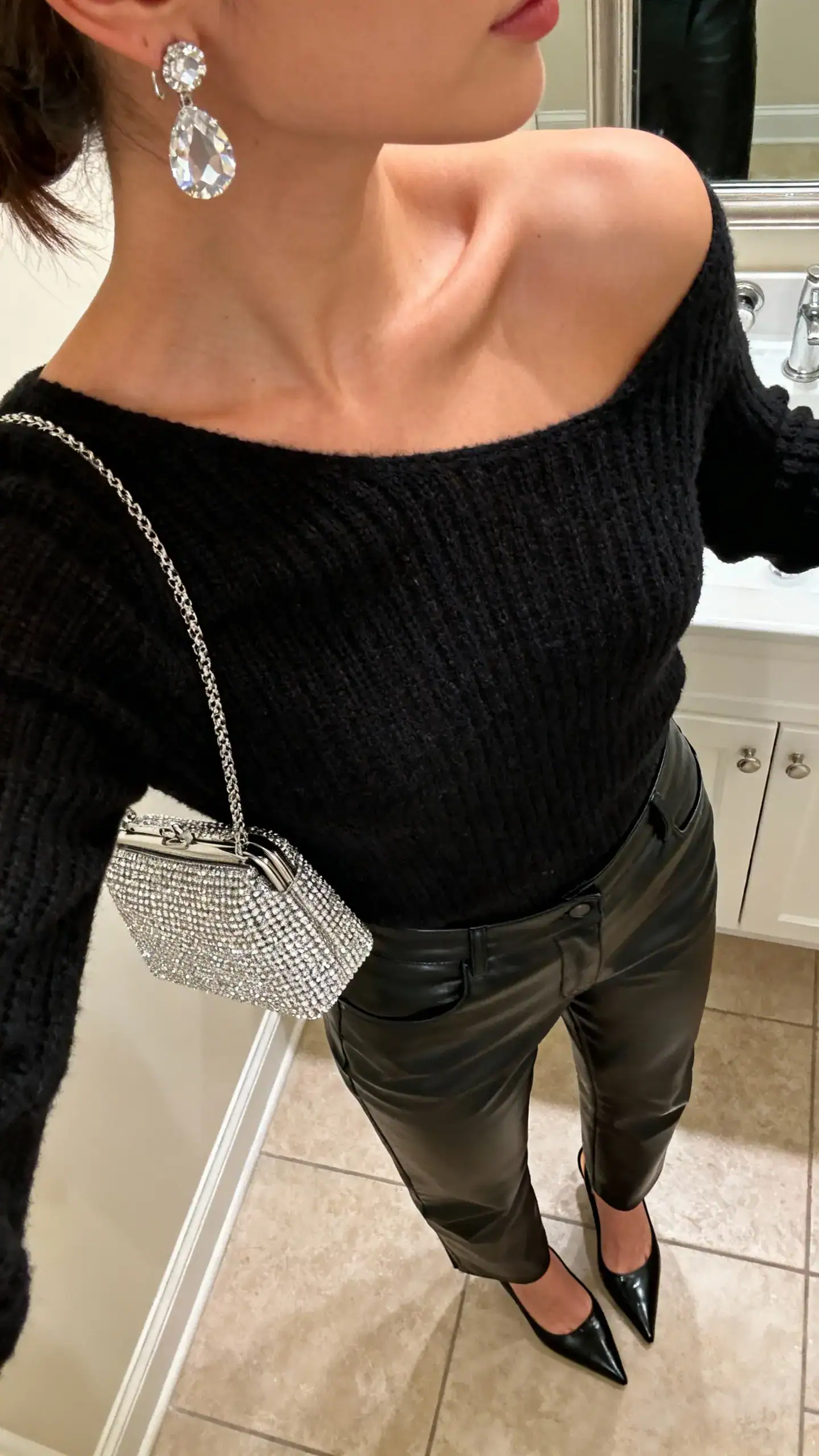 iPhone mirror selfie of a woman wearing an all-black look: leather pants, black knit top, black heels, with crystal earrings and a sparkling mini bag, face not visible, cropped at shoulders, bathroom mirror, warm indoor lighting, iPhone photo quality.