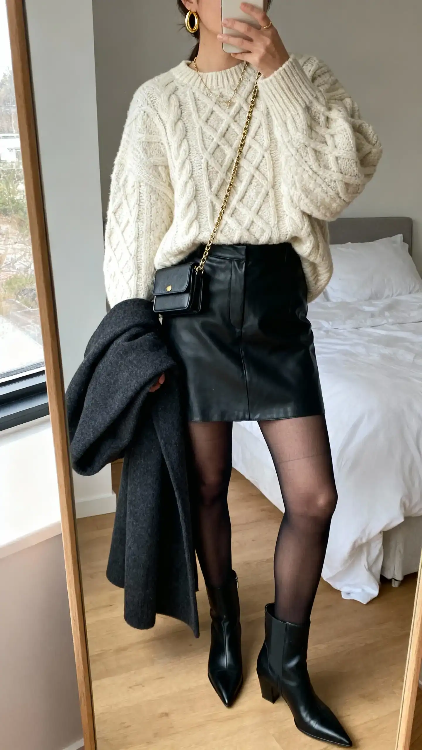 iPhone mirror selfie of a woman wearing an oversized cream cable-knit sweater, high-waisted black leather mini skirt, sheer black tights, black pointed-toe ankle boots, gold hoop earrings, delicate chain necklace, structured mini crossbody, and a long charcoal wool coat draped over arm; face not visible, cropped at neck, bedroom mirror, natural window light, iPhone photo quality.