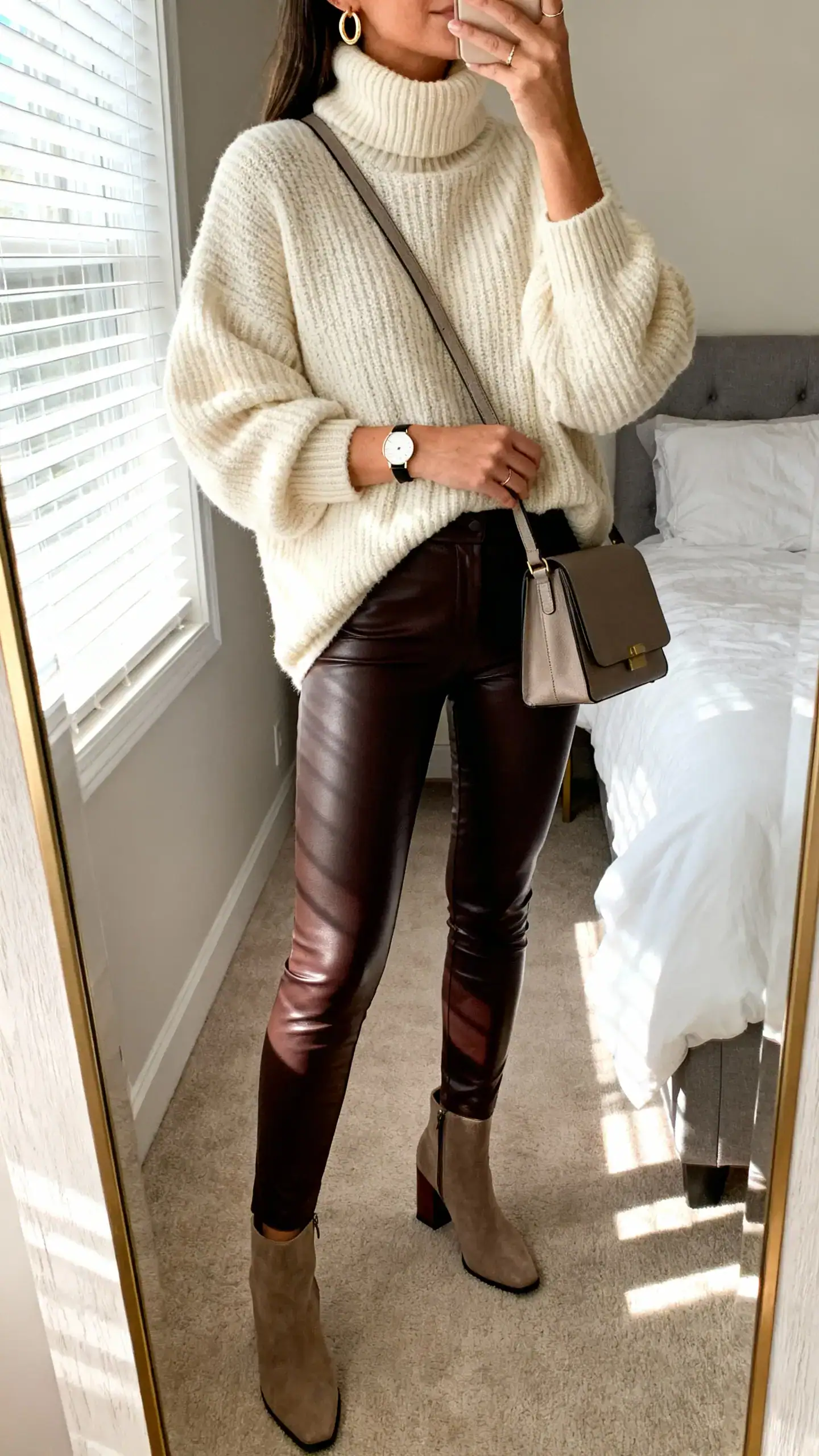iPhone mirror selfie of a woman wearing an oversized chunky turtleneck sweater in cream, high-waisted matte faux leather leggings, block-heel ankle boots, a structured crossbody bag, simple gold hoop earrings, and a slim watch, face not visible, cropped at neck, bedroom mirror with morning window light, iPhone photo quality.