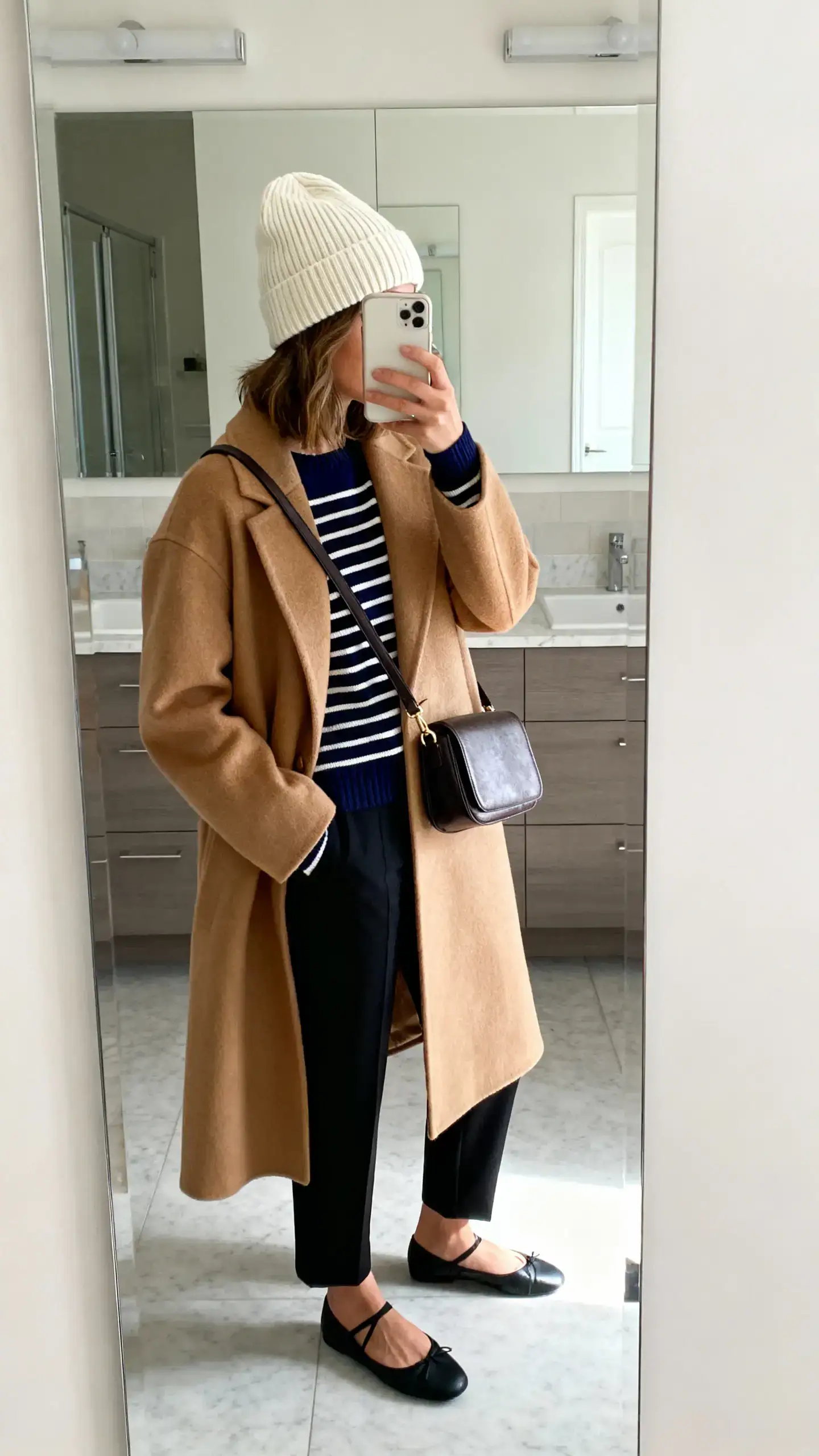 iPhone side-angle mirror selfie of a woman wearing a cream ribbed beanie, camel wool coat, navy striped Breton sweater, black cigarette trousers, ballet flats, and a small structured shoulder bag, face not visible, bathroom mirror, soft indoor lighting, iPhone photo quality.