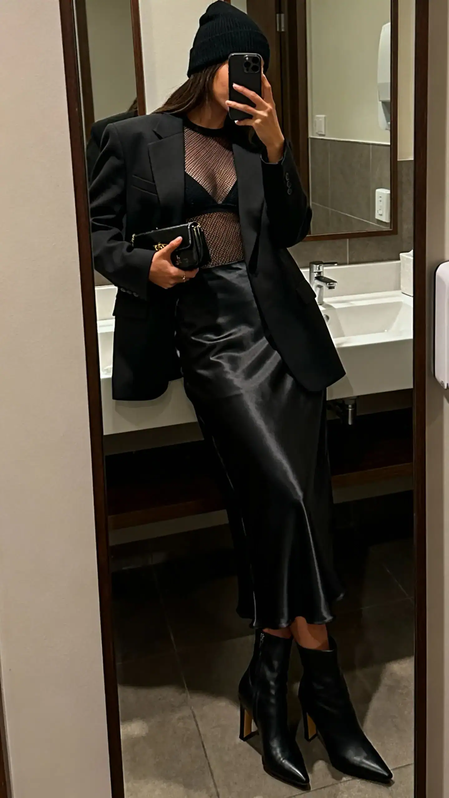 iPhone side-angle mirror selfie of a woman wearing a black beanie, long satin slip skirt, fitted mesh top under a tailored blazer, heeled ankle boots, and a small clutch, face not visible, cropped at neck, bathroom mirror, dim warm lighting, iPhone photo quality.