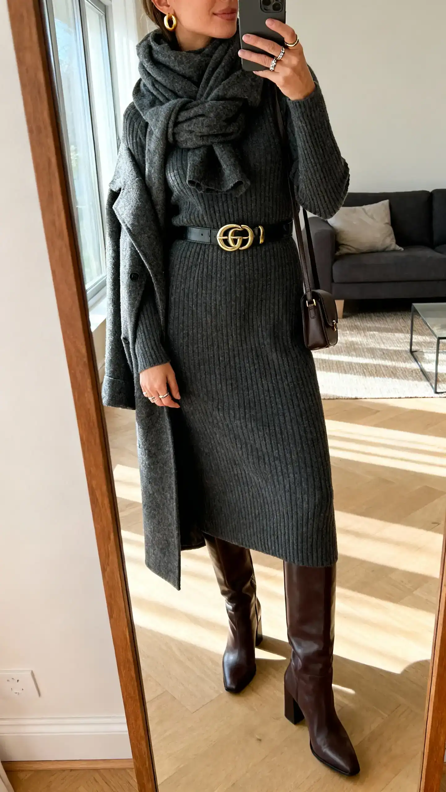 iPhone side-angle mirror selfie of a woman wearing a ribbed charcoal midi sweater dress, knee-high leather boots with block heels, chunky scarf, statement waist belt, gold huggie earrings, stacking rings, longline wool coat, and a small crossbody; face not visible, living room mirror, afternoon window light, iPhone photo quality.