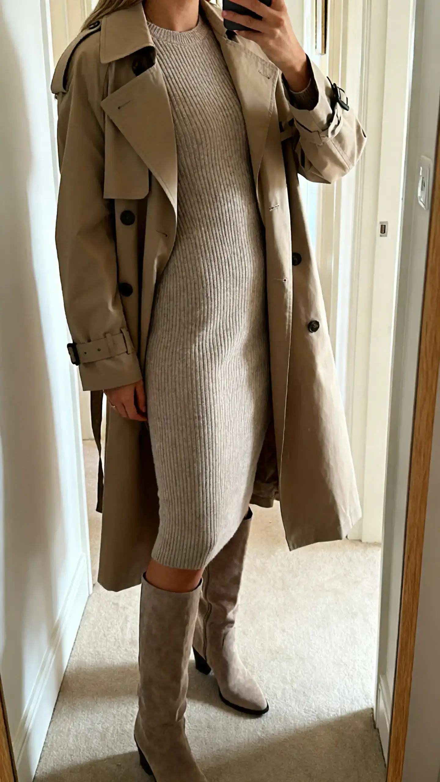 iPhone side-angle selfie of a woman wearing a bodycon knit dress with tall boots and a classic trench coat, face not visible, cropped at shoulders, hallway mirror, natural morning light, iPhone photo quality.