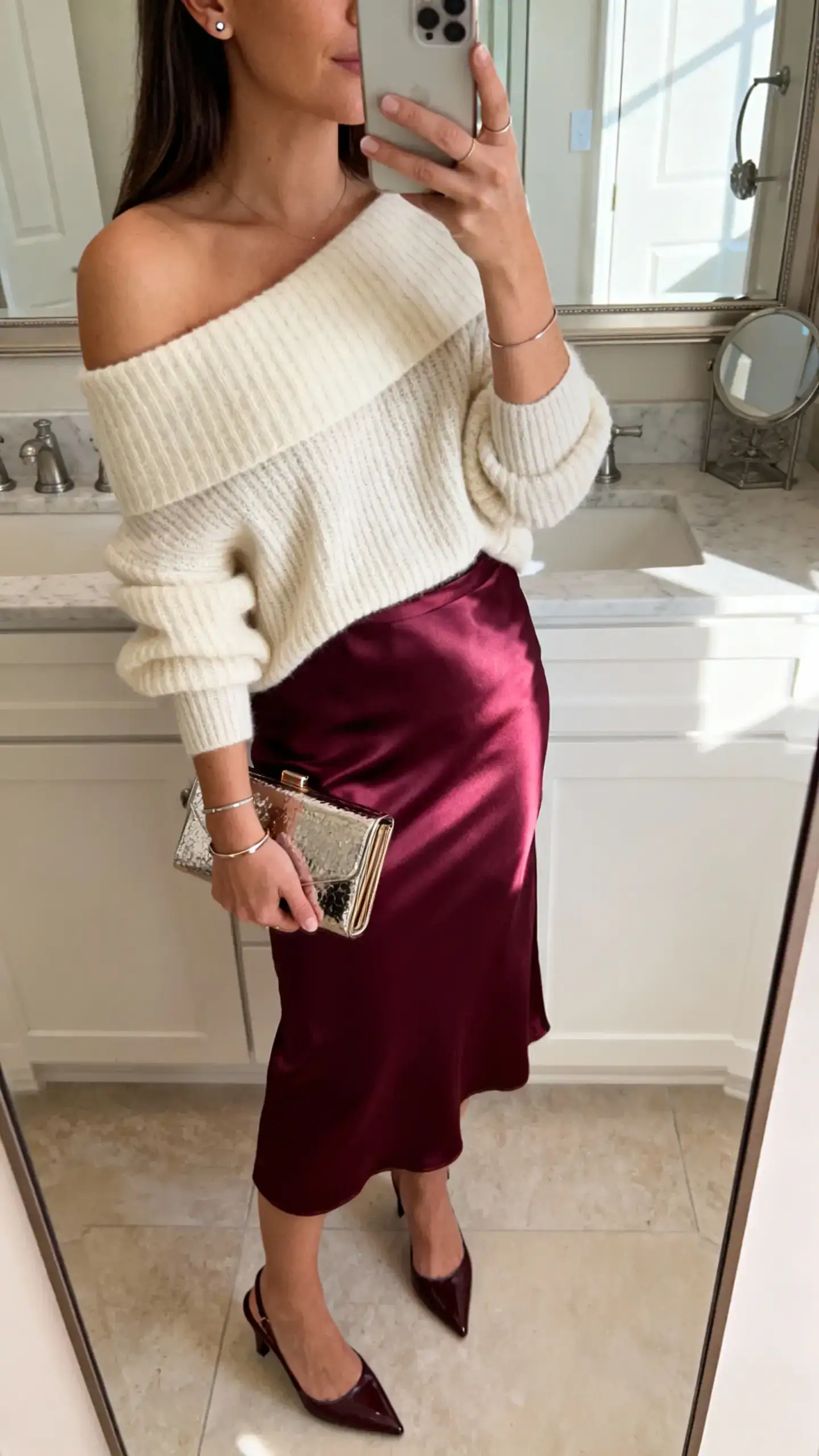 iPhone mirror selfie of a woman in a berry satin midi skirt, off-shoulder cream knit, sleek slingbacks, minimal stud earrings, slim bracelet, and a compact clutch with metallic detailing; face not visible, bathroom mirror, bright morning light, iPhone photo quality.
