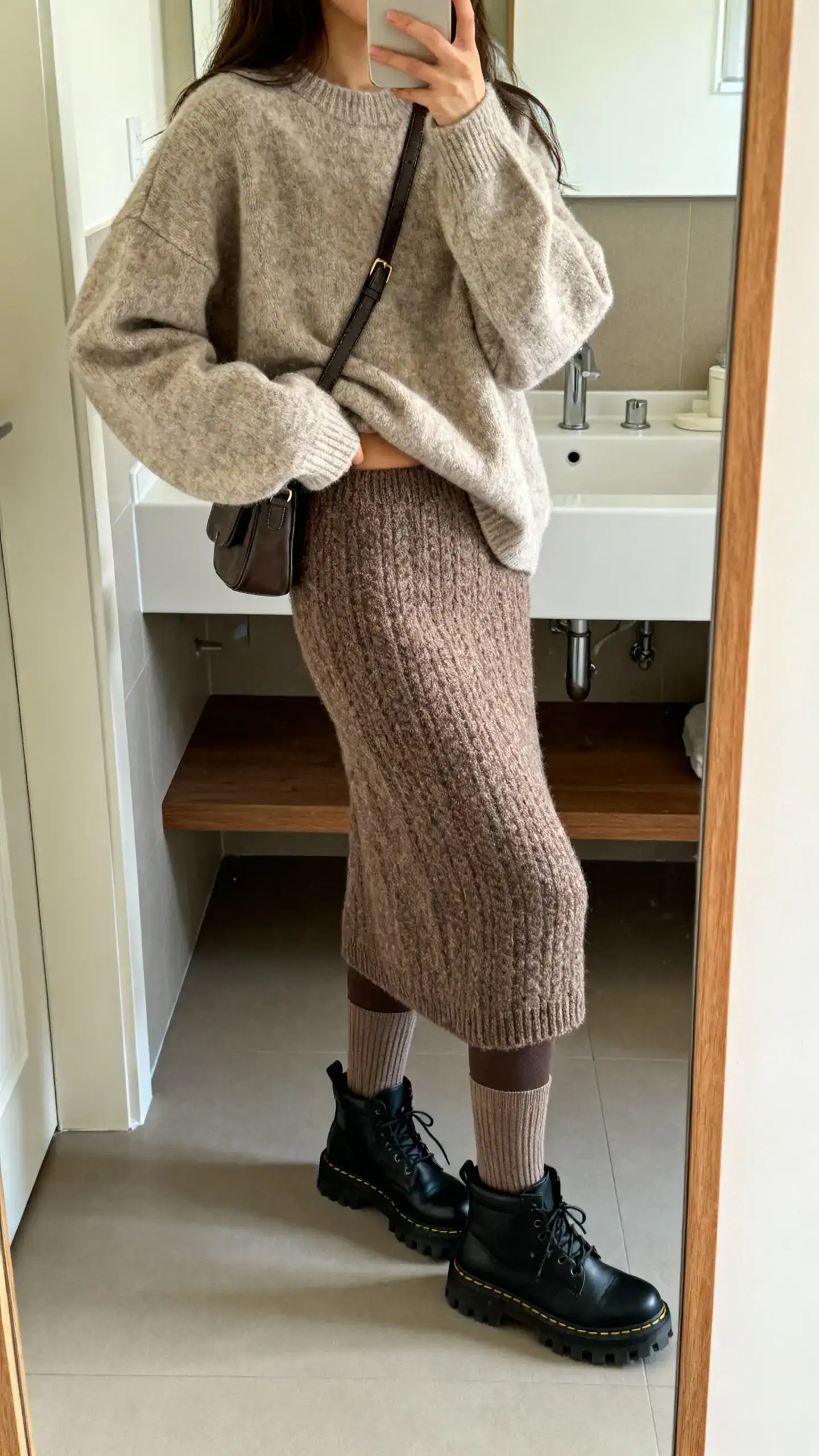 iPhone mirror selfie of a woman in a cozy oversized sweater half-tucked into a warm knit midi skirt, thermal tights, lug-sole boots, and a crossbody bag, face not visible, side angle in bathroom mirror, soft indoor lighting, iPhone photo quality.