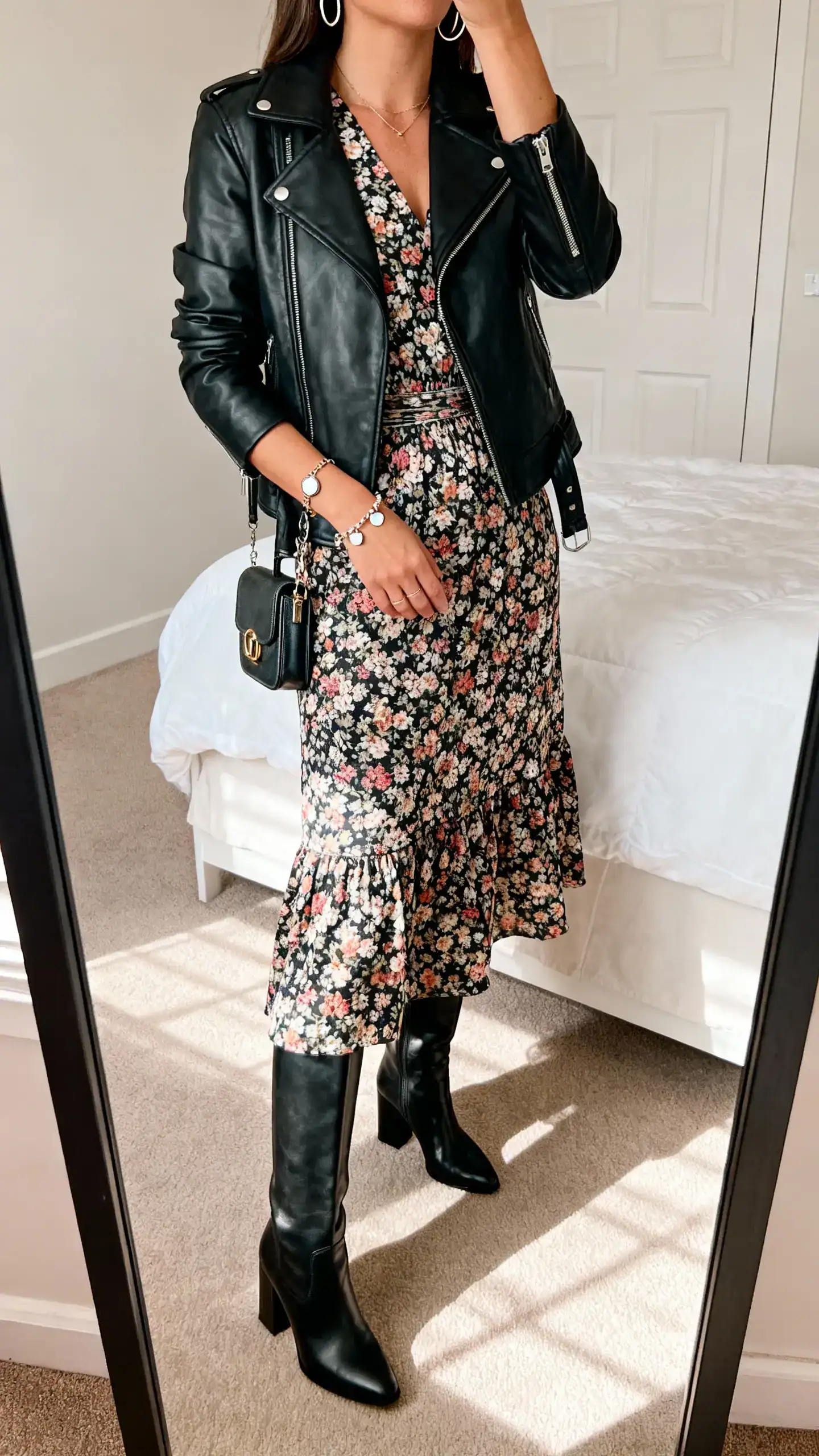 iPhone mirror selfie of a woman in a floral midi dress with a defined waist, black leather moto jacket, knee-high block-heel boots, delicate hoops, charm bracelet, and a mini satchel; face not visible, cropped at shoulders, bedroom mirror, afternoon natural light, iPhone photo quality.
