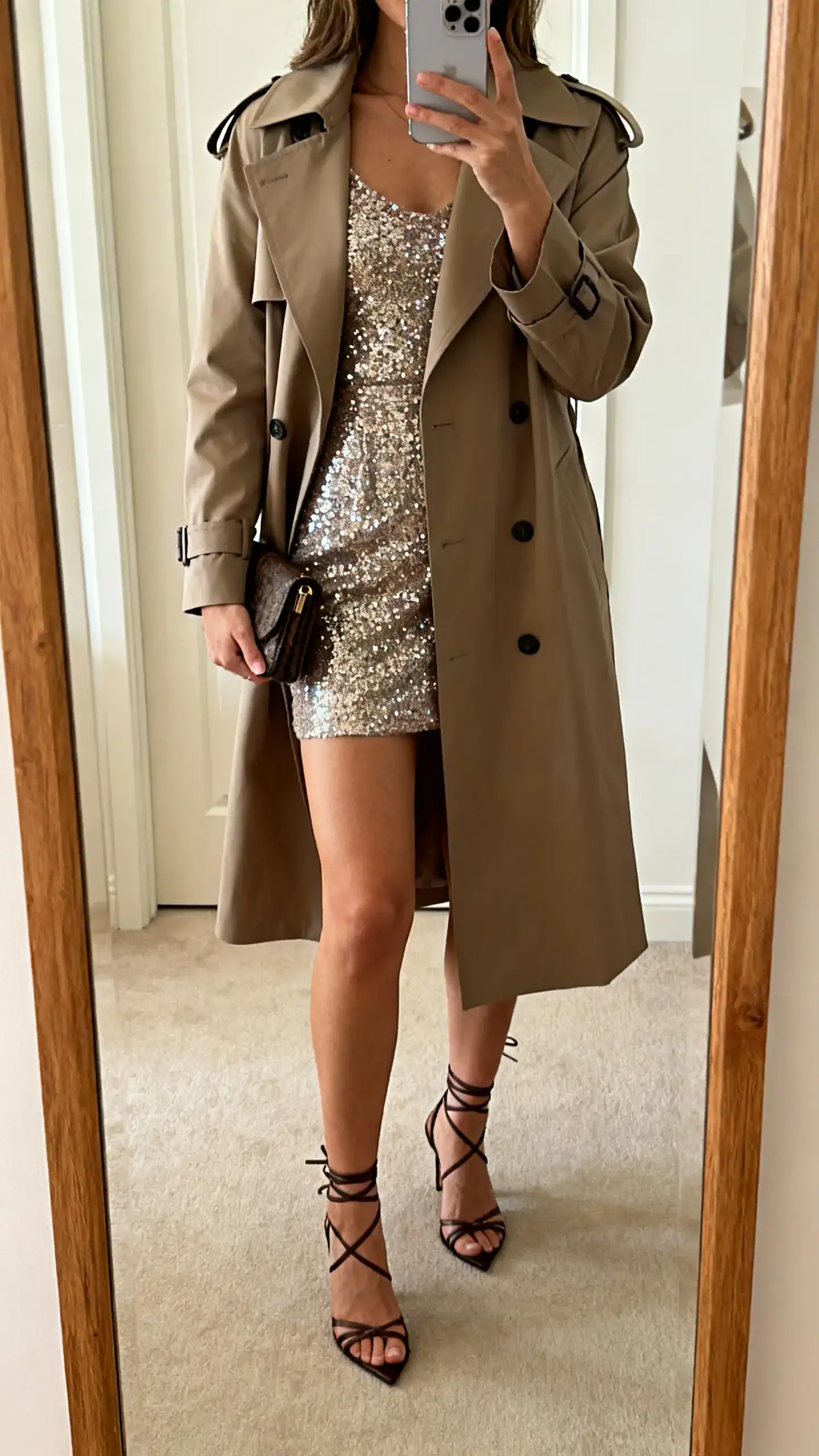 iPhone mirror selfie of a woman in a sparkly mini dress layered with a tailored trench coat, strappy heels, and a small clutch, face not visible, cropped at shoulders, full-length mirror, warm indoor lighting, iPhone photo quality.