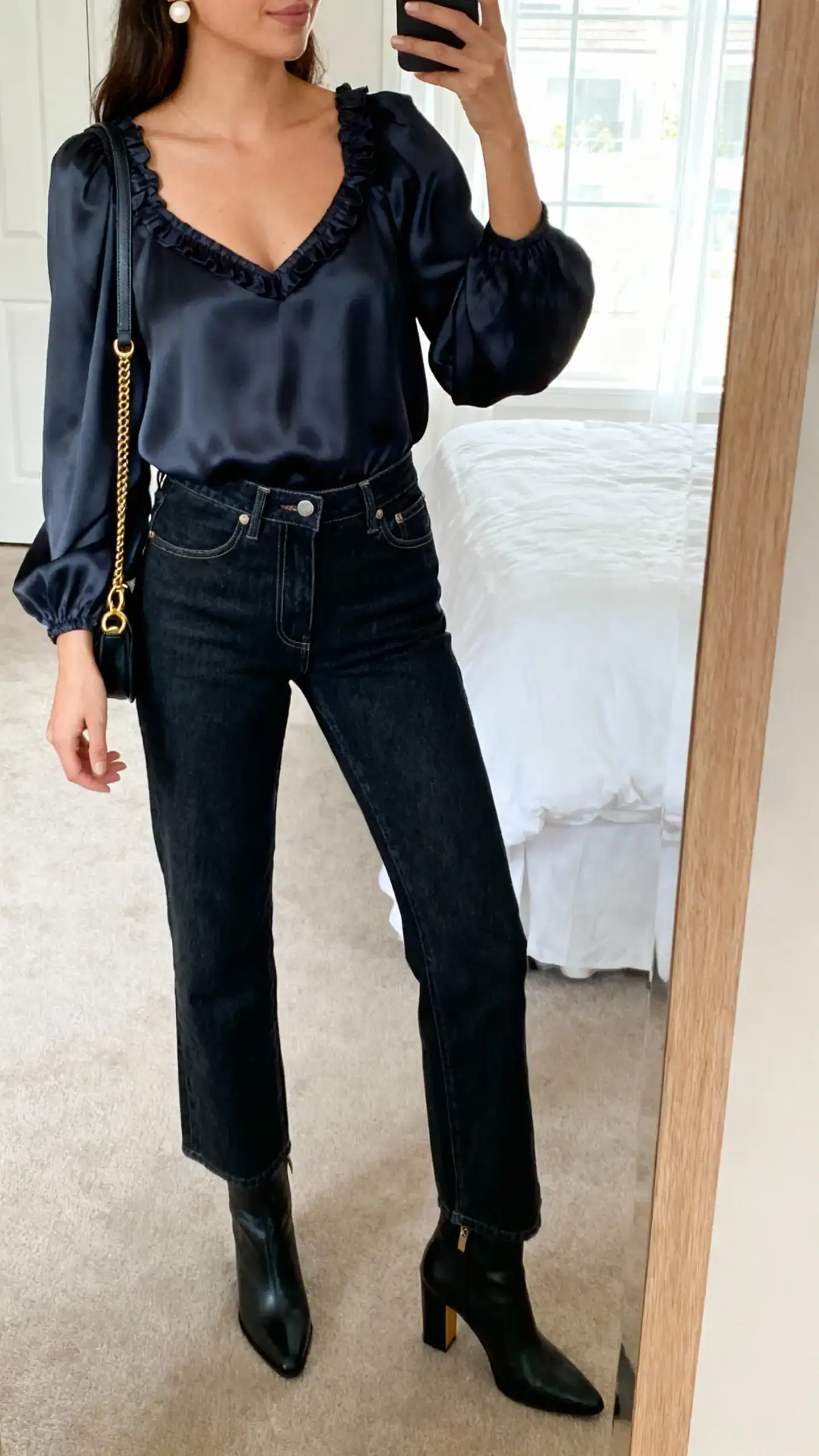 iPhone mirror selfie of a woman in dark-wash high-waisted straight-leg jeans, a romantic silky blouse with a sweetheart neckline and subtle ruffles, heeled ankle boots, pearl studs, and a crossbody bag with gold hardware; face not visible, cropped at shoulders, bedroom mirror, daylight, iPhone photo quality.