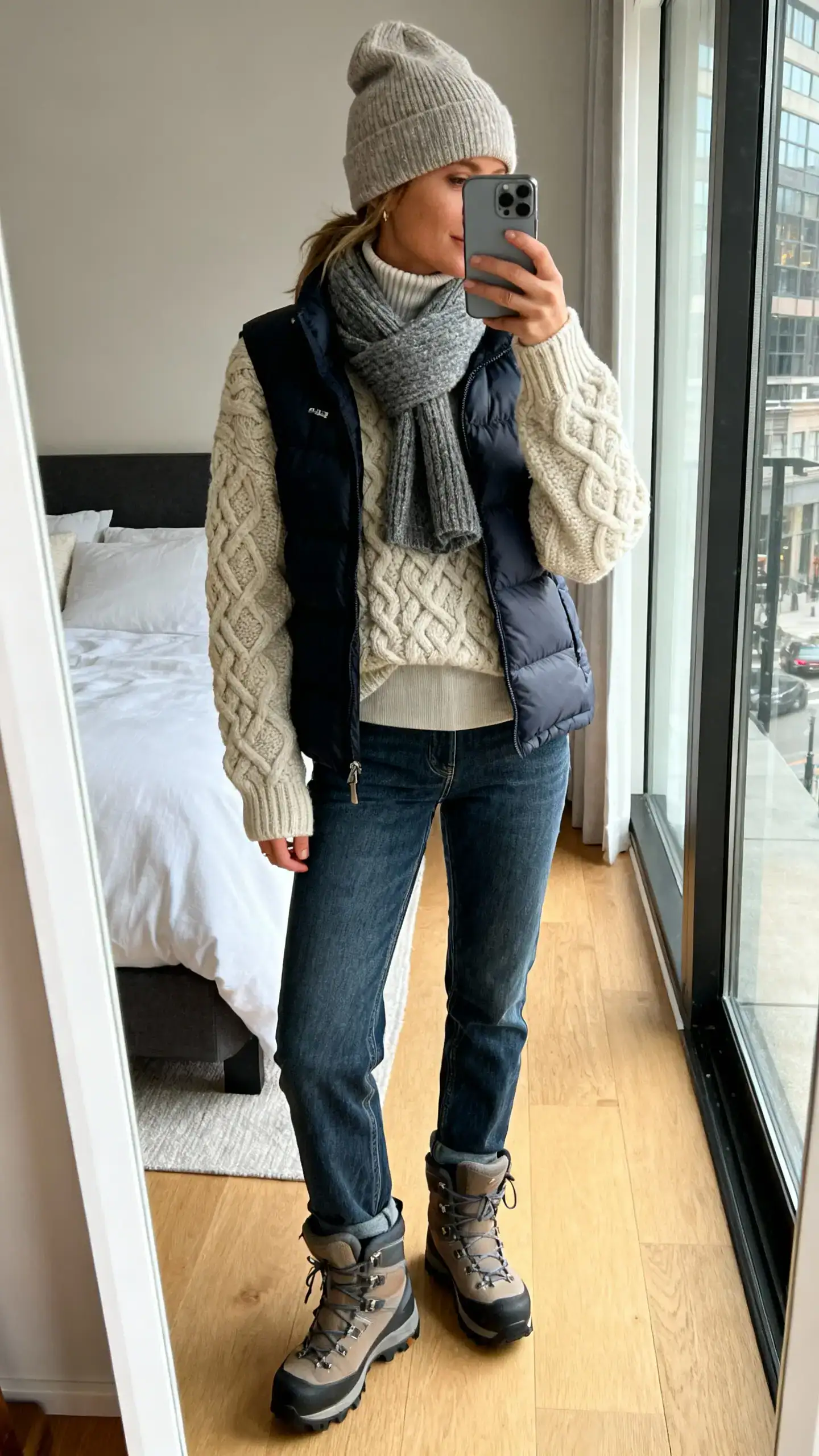 iPhone mirror selfie of a woman layered in an apr&egrave;s-ski city look: chunky cable knit sweater over a thin turtleneck, beanie, wool scarf, puffer vest, straight jeans, and hiking-inspired boots, face not visible, cropped at neck, bedroom mirror, morning light, iPhone photo quality.