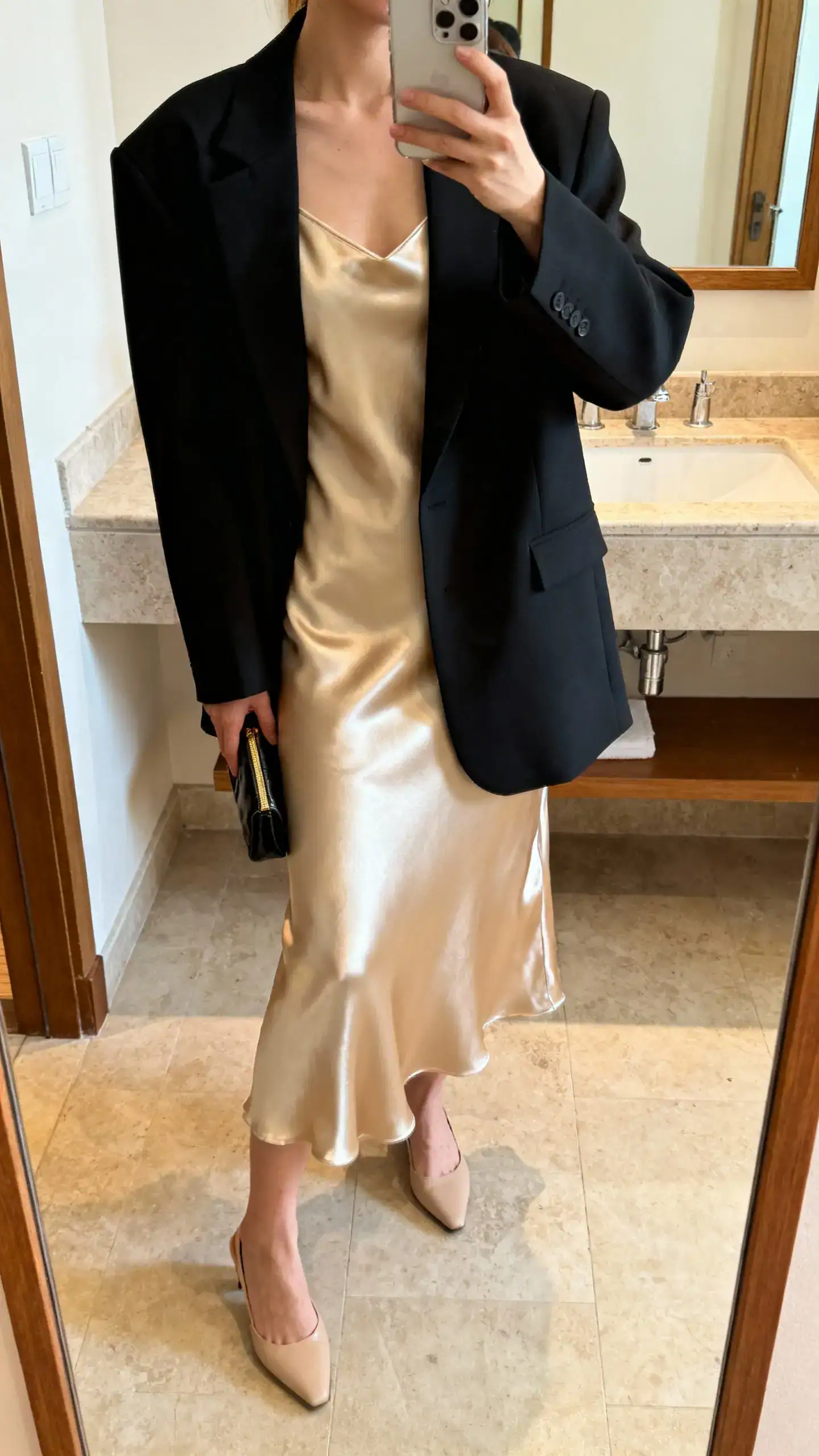 iPhone mirror selfie of a woman wearing a champagne satin slip dress under a boxy black blazer with minimalist heels and a small clutch, face not visible, bathroom mirror, warm indoor lighting, iPhone photo quality.