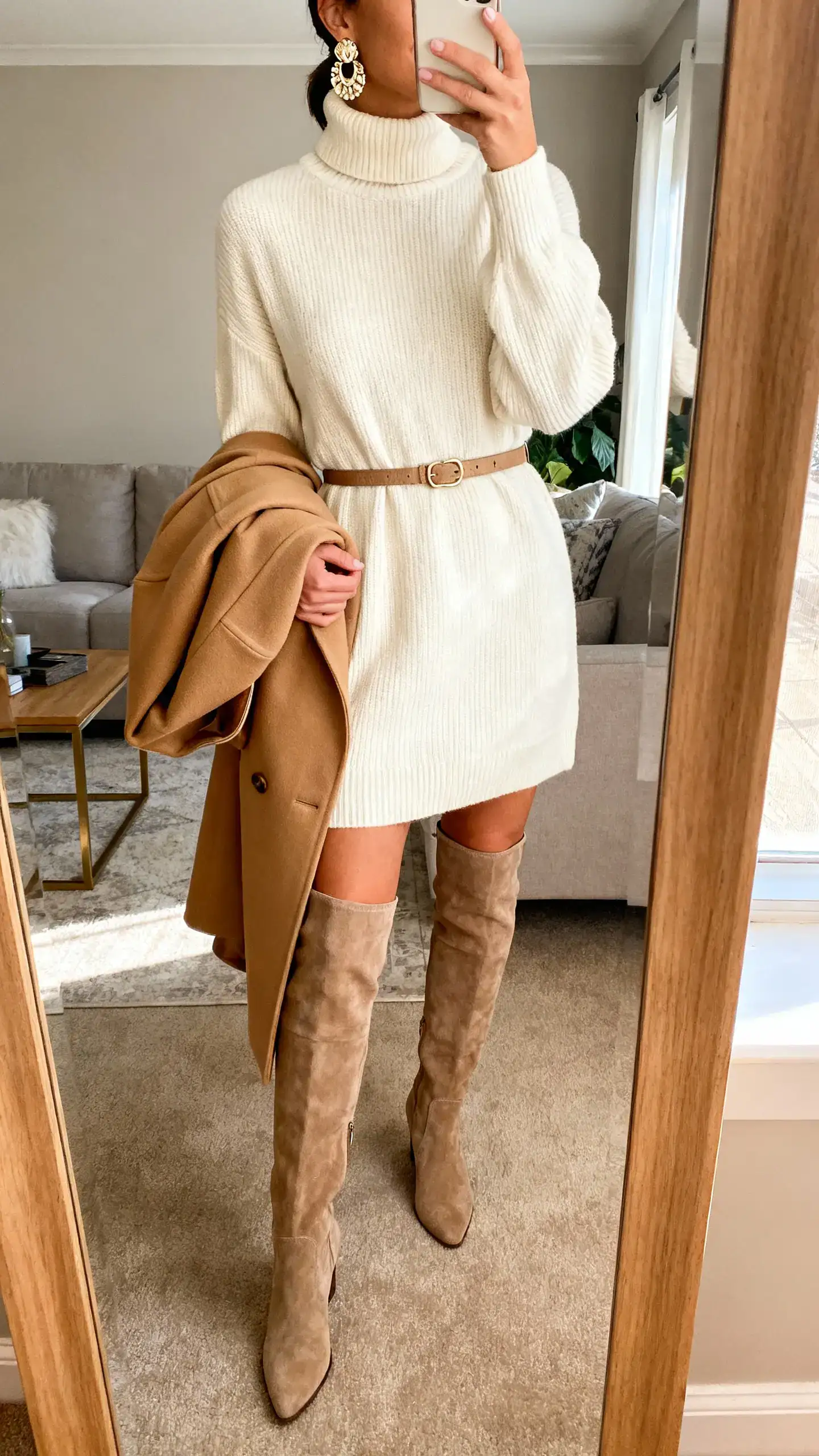 iPhone mirror selfie of a woman wearing a cream turtleneck sweater dress with an optional waist belt, over-the-knee boots, statement earrings, and a camel structured coat draped over her arm; face not visible, cropped at neck, living room mirror, warm indoor lighting, iPhone photo quality.
