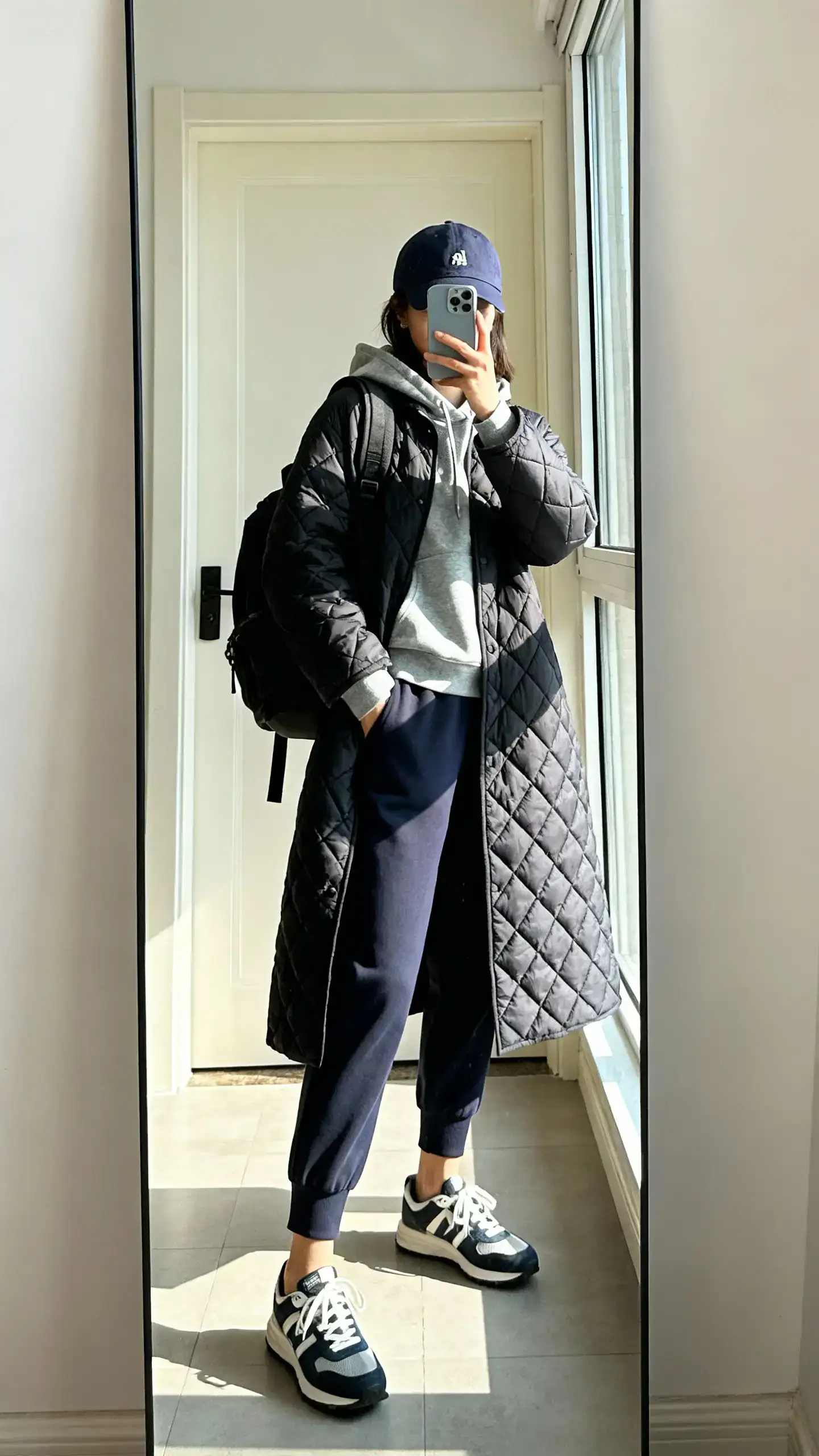 iPhone mirror selfie of a woman wearing a quilted coat, hoodie, tapered joggers, classic sneakers, baseball cap, and a backpack, face not visible, full-length mirror by the door, natural window light, iPhone photo quality.