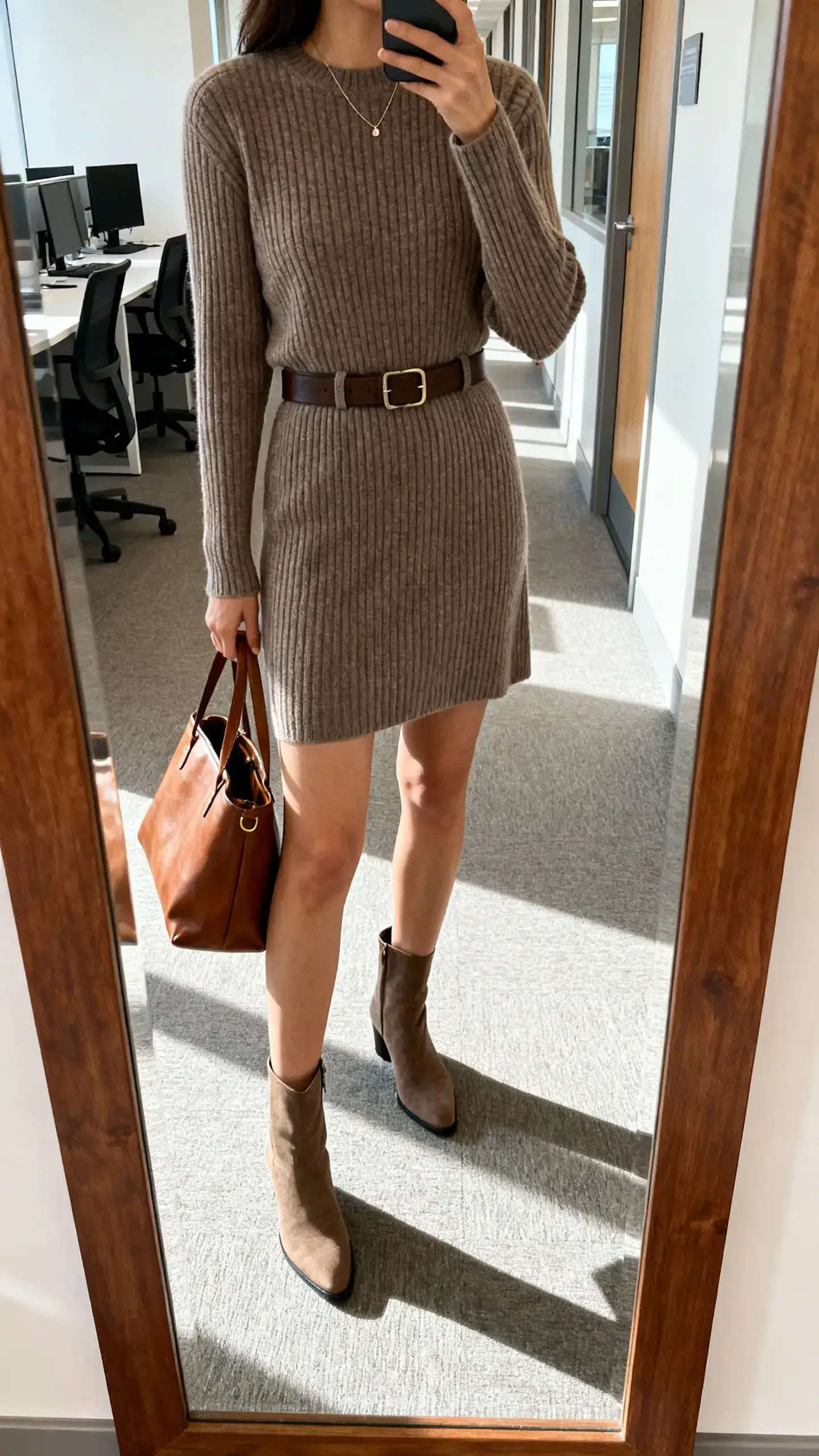 iPhone mirror selfie of a woman wearing a ribbed knit dress with a belt, slim knee boots, minimal necklace, and a leather tote, face not visible, cropped at shoulders, office hallway mirror, natural daylight, iPhone photo quality.
