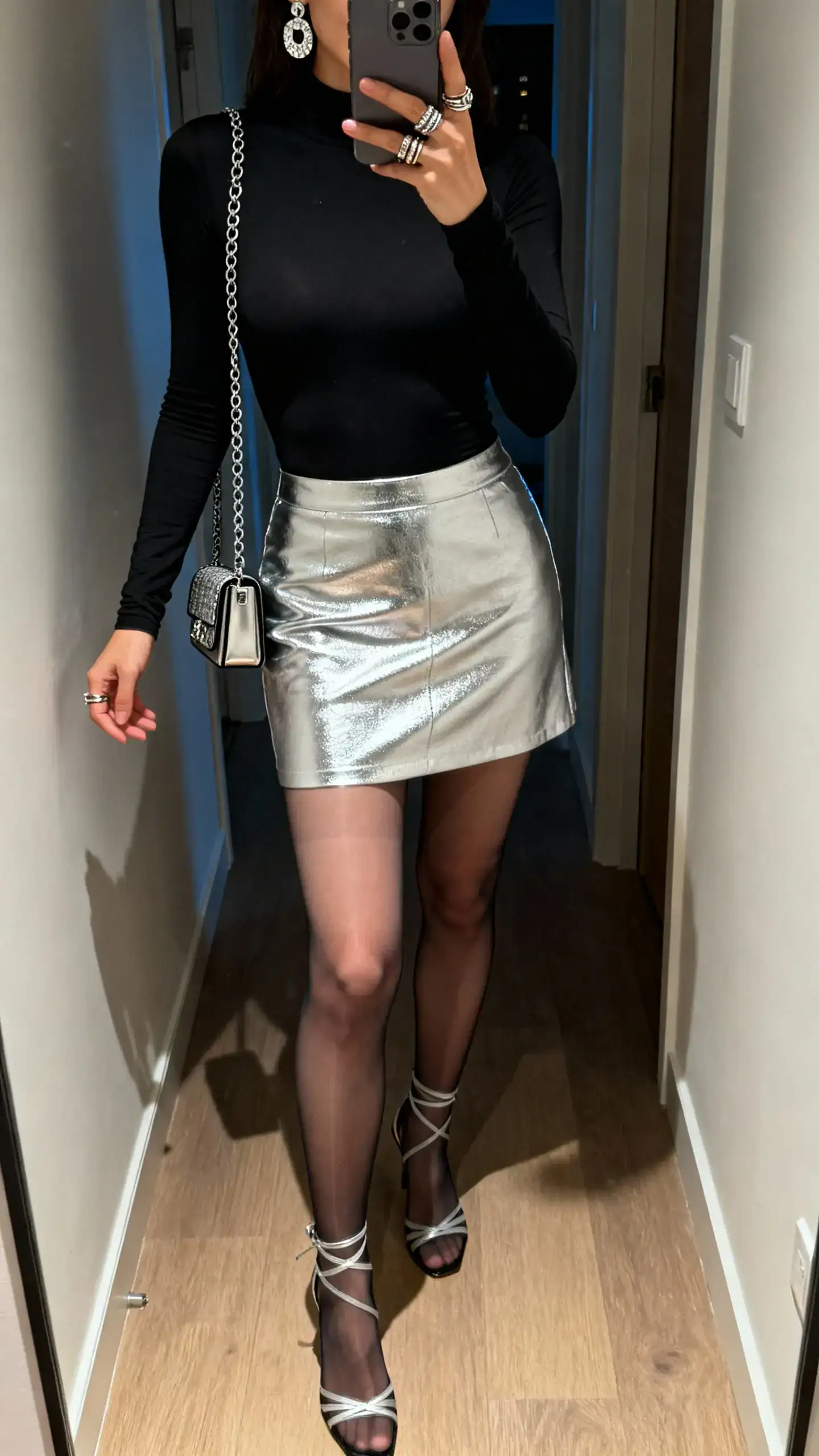 iPhone mirror selfie of a woman wearing a silver metallic mini skirt, fitted black long-sleeve bodysuit, sheer tights, strappy heels, statement earrings, stackable rings, and a compact chain-strap shoulder bag; face not visible, hallway mirror, evening indoor light, iPhone photo quality.