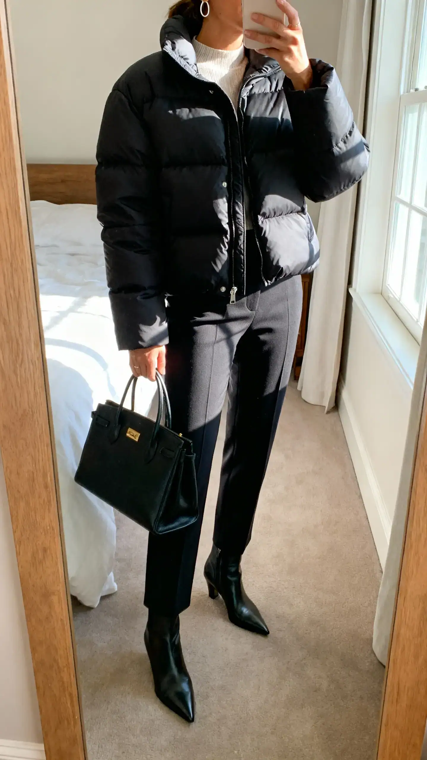 iPhone mirror selfie of a woman wearing a sleek elevated puffer jacket, tailored trousers, pointed ankle boots, structured tote, and minimal hoops, face not visible, cropped at neck, bedroom mirror, morning window light, iPhone photo quality.