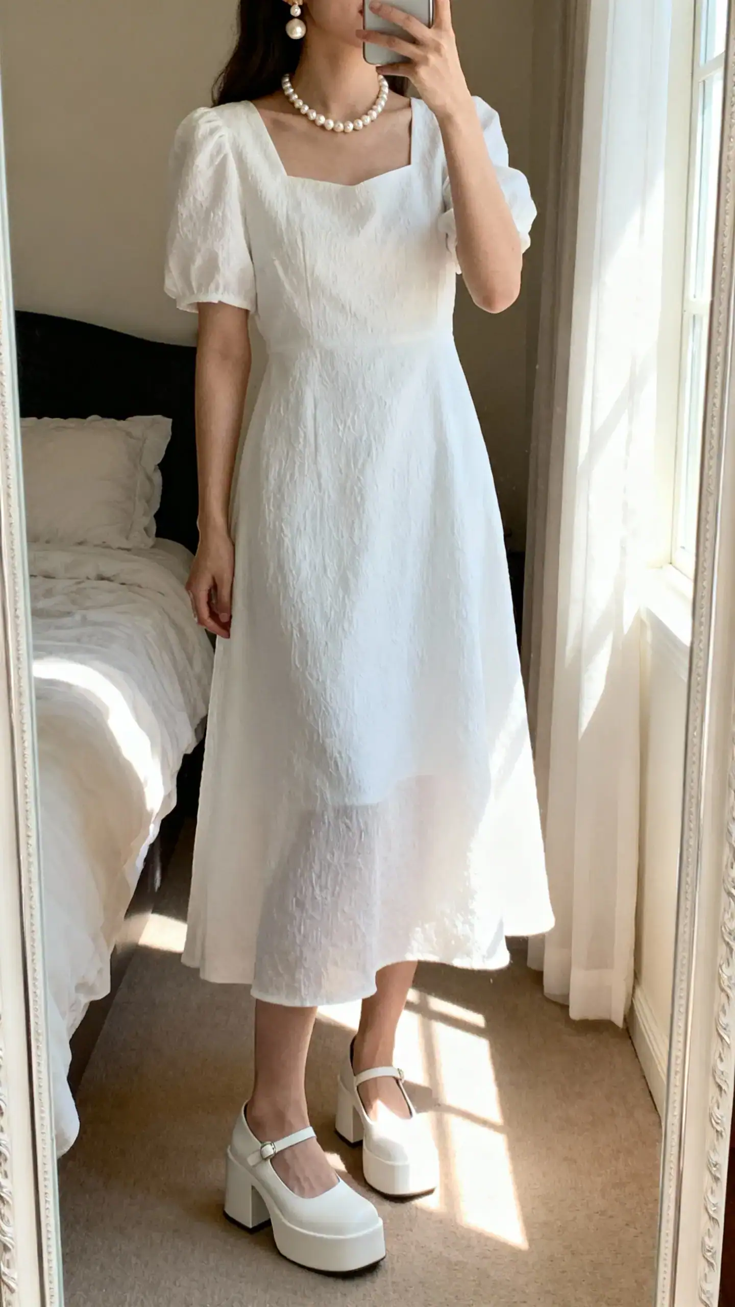 iPhone mirror selfie of a woman wearing a white dress with pearl necklace, pearl earrings, and white platform heels, face not visible, cropped at neck, bedroom mirror, soft morning window light, iPhone photo quality.