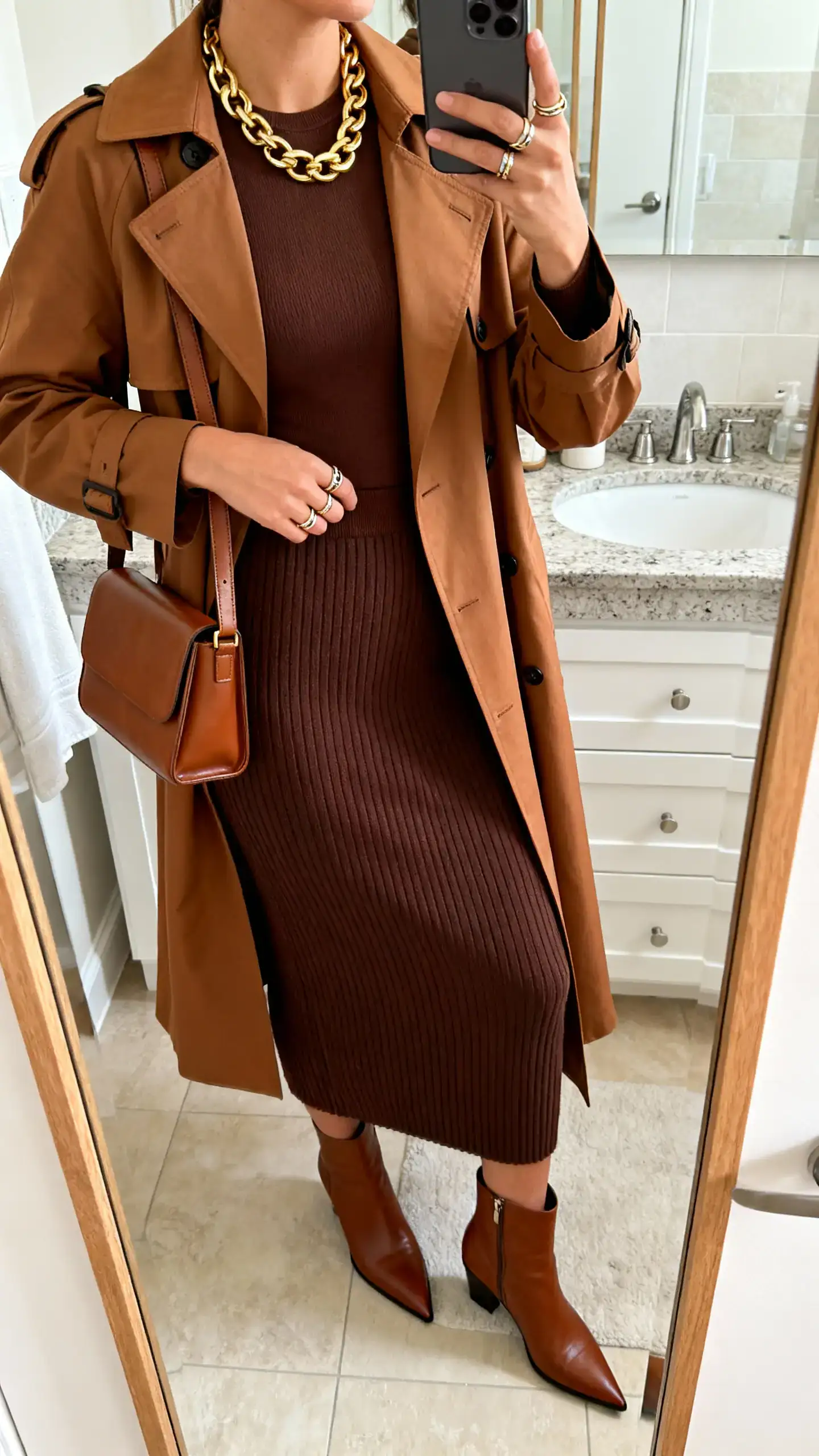 iPhone side-angle mirror selfie of a woman in a chocolate rib-knit midi skirt with matching fitted top, caramel trench coat, brown pointed-toe ankle boots, chunky gold chain necklace, stacked rings, and a structured brown shoulder bag; face not visible, bathroom mirror, morning indoor light, iPhone photo quality.