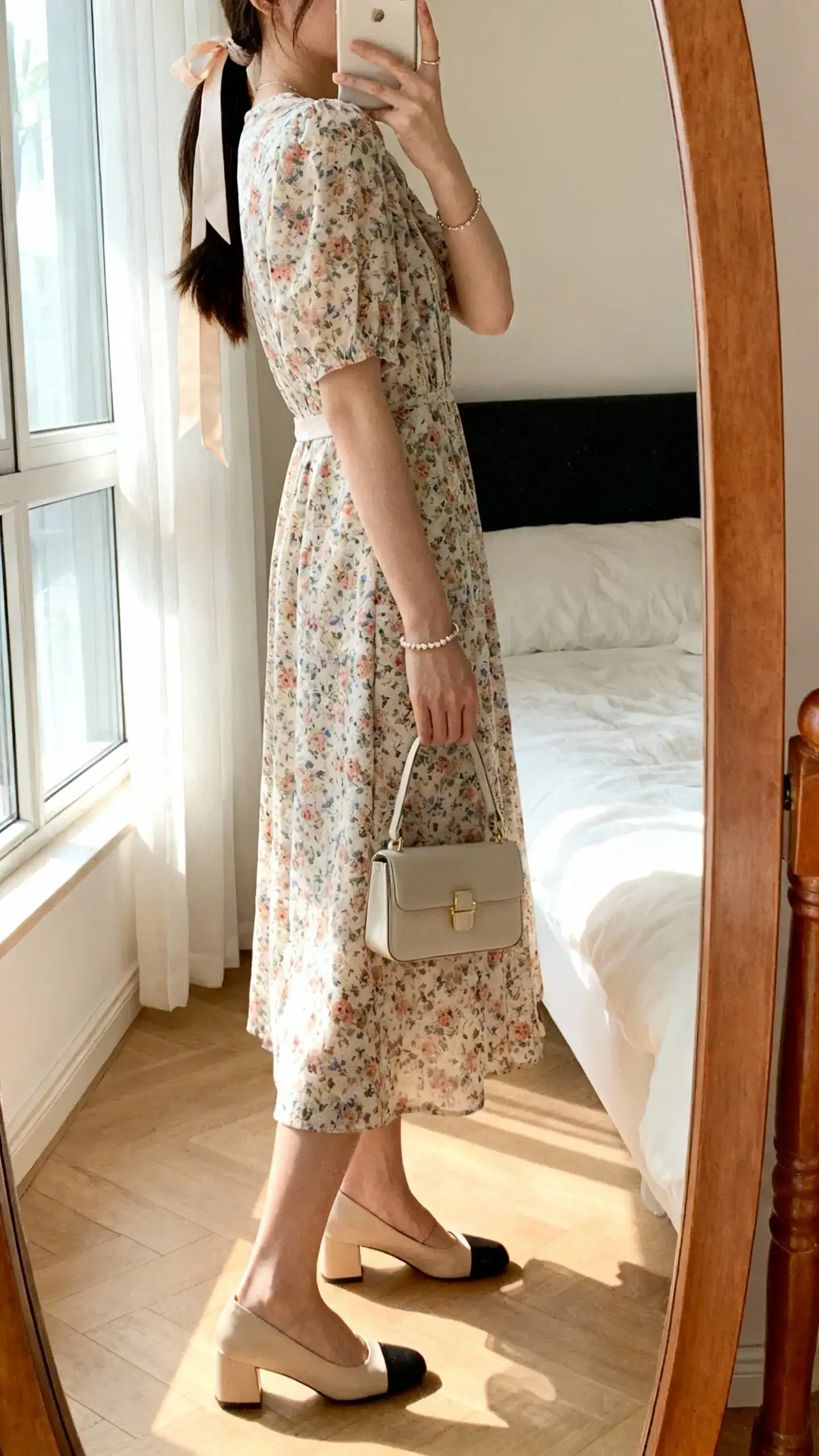 iPhone side-angle mirror selfie of a woman in a floral midi dress with block heels and a ribbon-tied low ponytail, delicate bracelet and mini bag, face not visible, bedroom mirror, afternoon window light, iPhone photo quality.