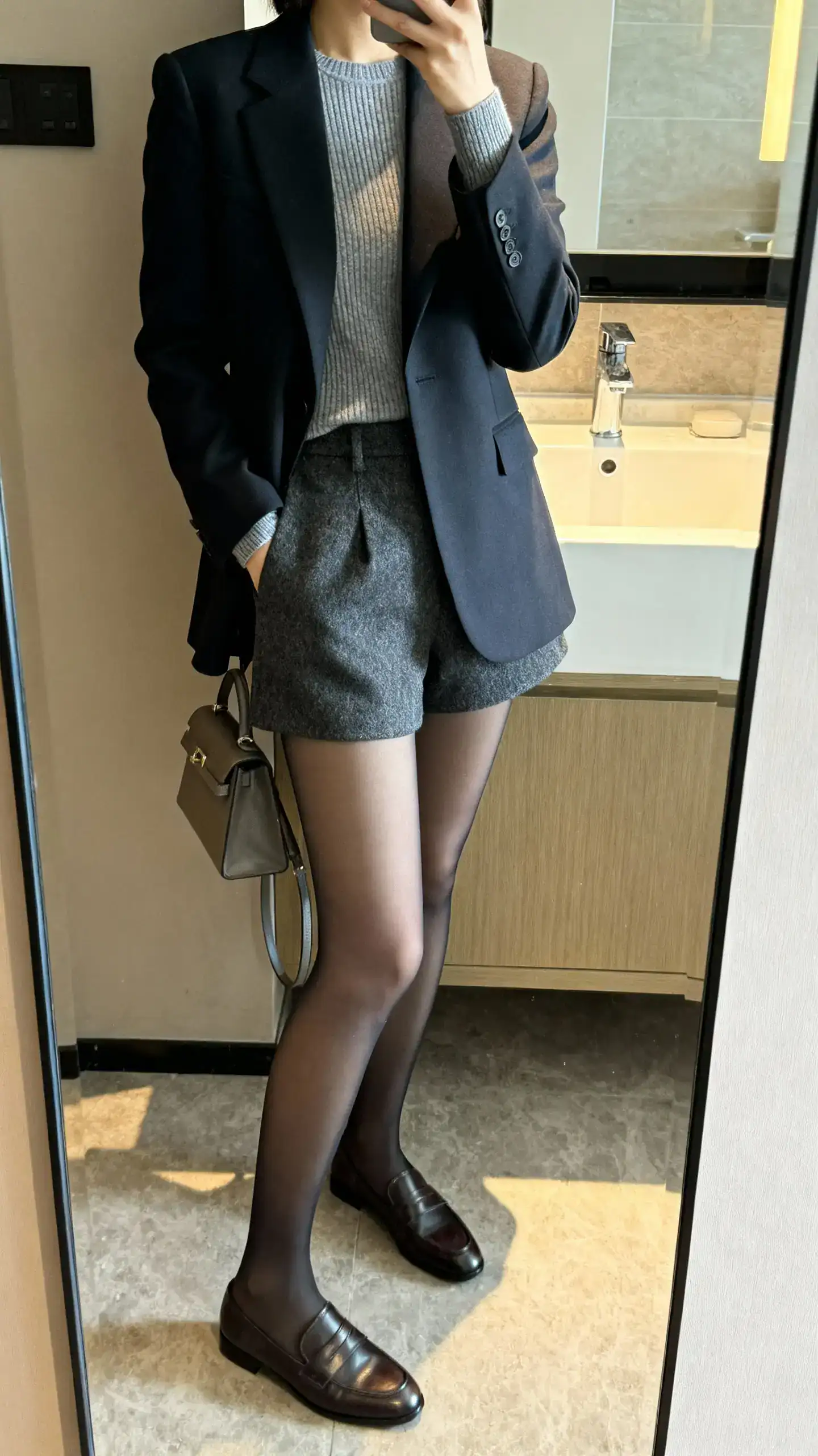 iPhone side-angle mirror selfie of a woman in opaque tights with tailored wool shorts, a fitted blazer over a fine knit, loafers, and a small structured bag, face not visible, cropped at neck, bathroom mirror, indoor lighting, iPhone photo quality.